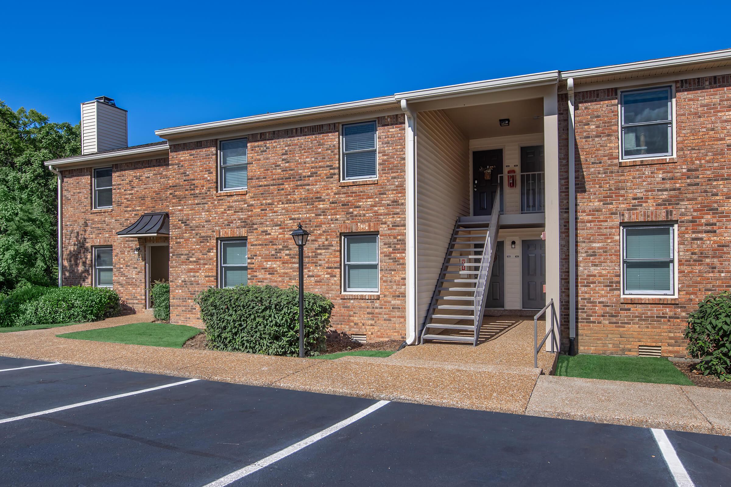 A two-story apartment building with a brick facade. The building features a set of stairs leading to the second floor, with doors on both levels. There are green shrubs and a lamp post in front, along with a paved parking area. The sky is clear and blue, indicating a sunny day.