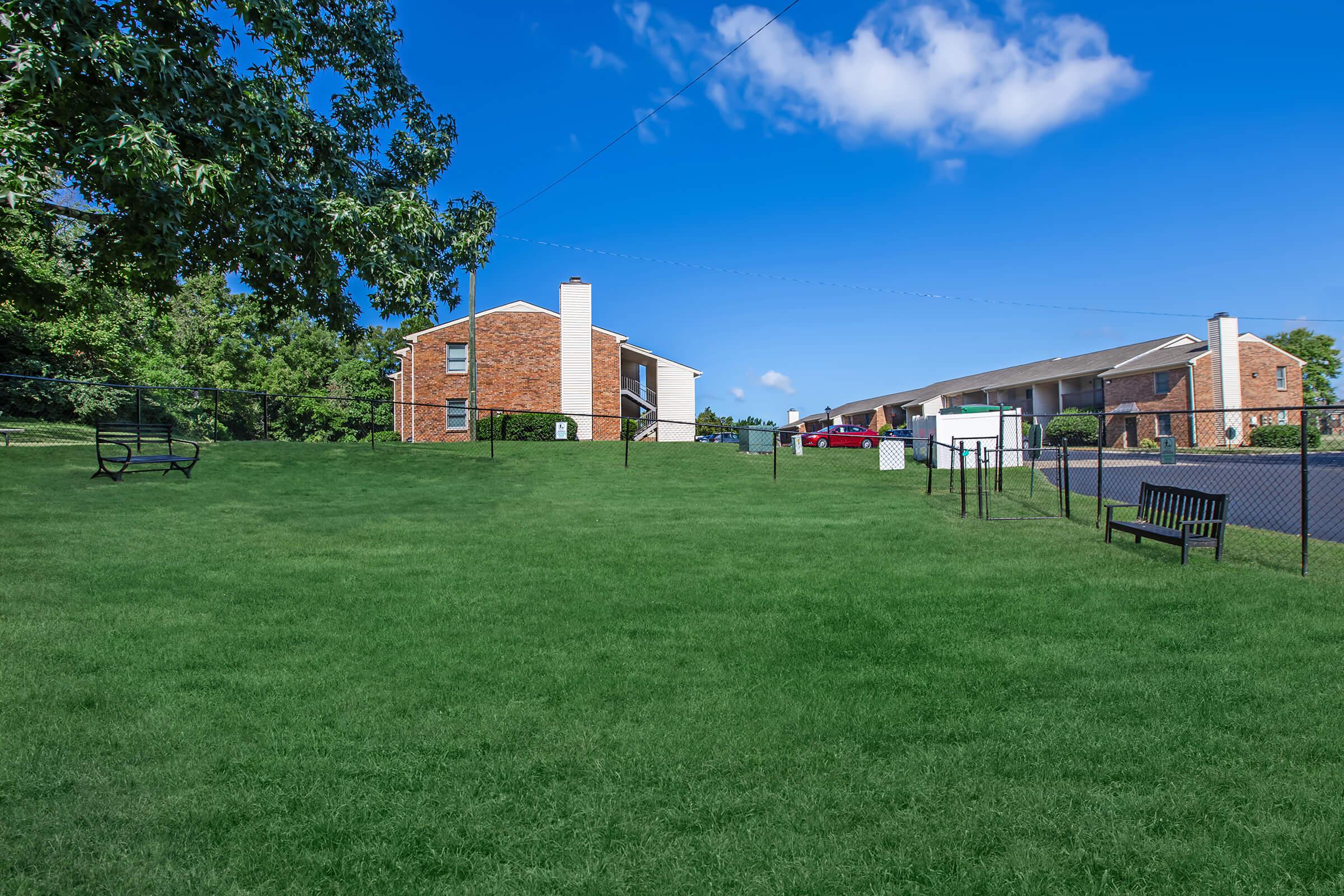 A green grassy area in a residential neighborhood featuring two brick apartment buildings. There is a wooden bench and a swing set on the grass, surrounded by a fence, with a clear blue sky and a few white clouds above.