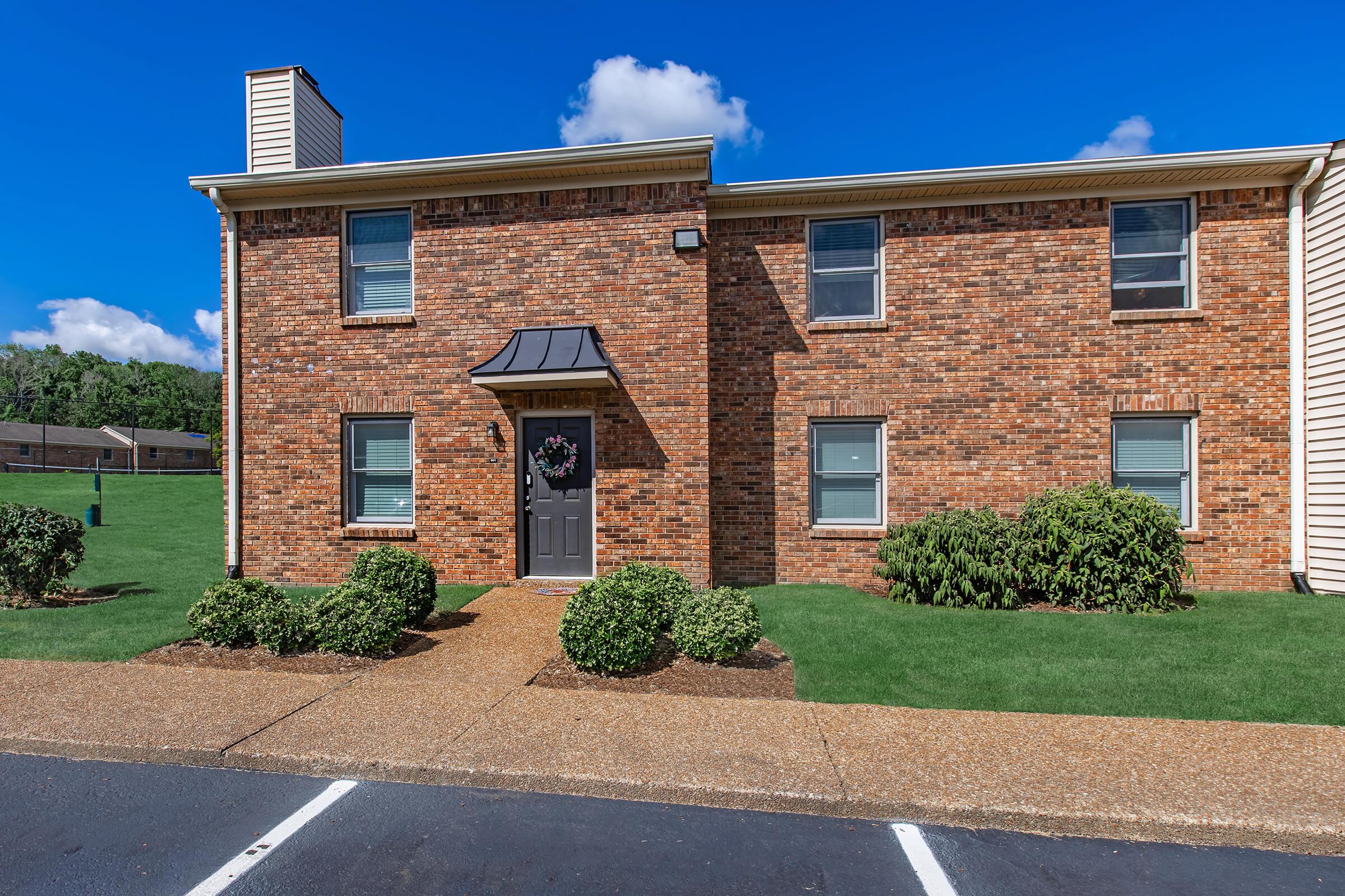 A brick apartment building with a dark front door, flanked by green bushes. The path leading to the door is made of gravel, and the area is well-kept with grass. The sky is bright blue with a few clouds in the background.