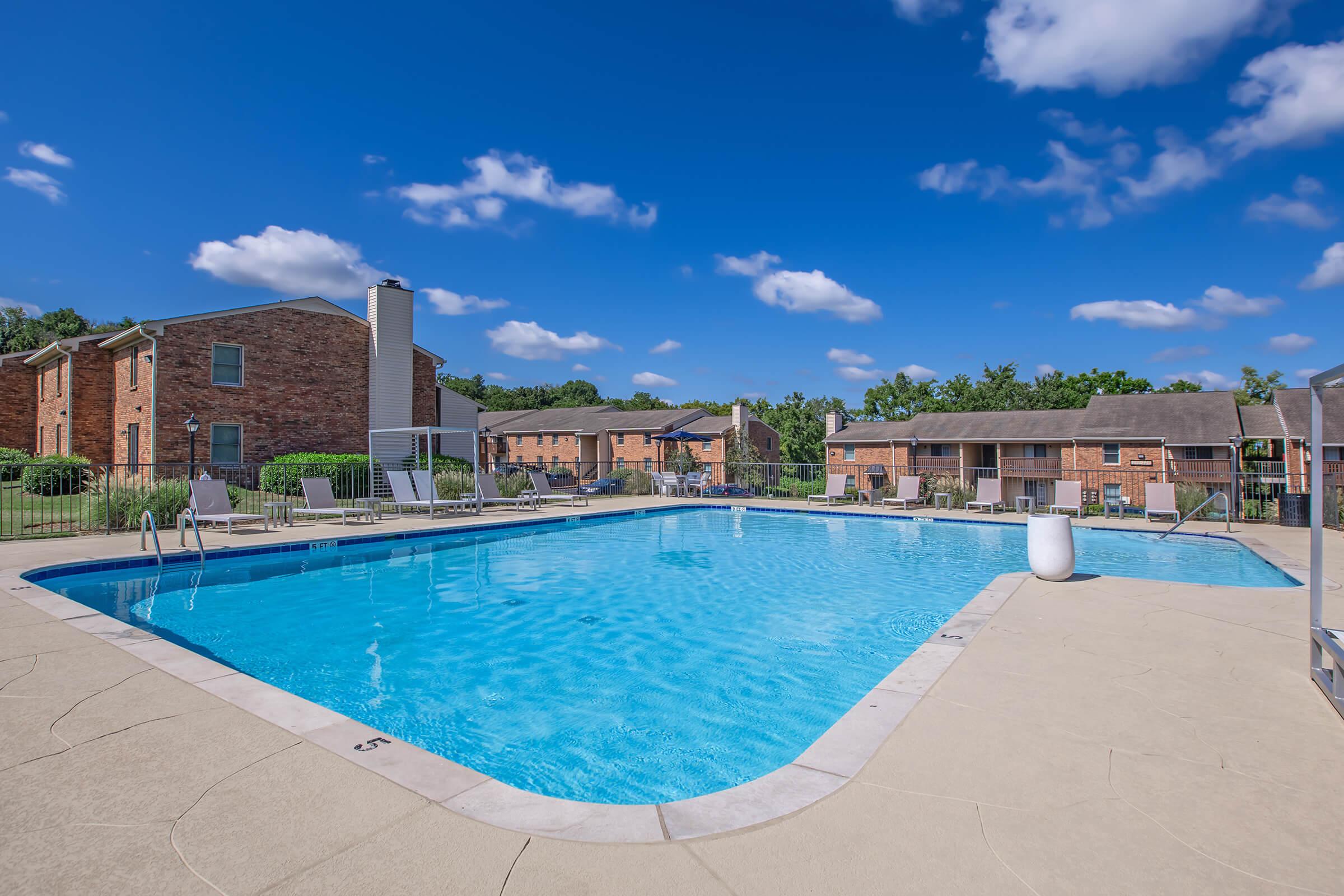 A clear blue swimming pool surrounded by a concrete deck, with lounge chairs and tables arranged nearby. In the background, brick apartment buildings are visible against a bright blue sky with fluffy white clouds. Green trees line the area, creating a serene outdoor atmosphere.