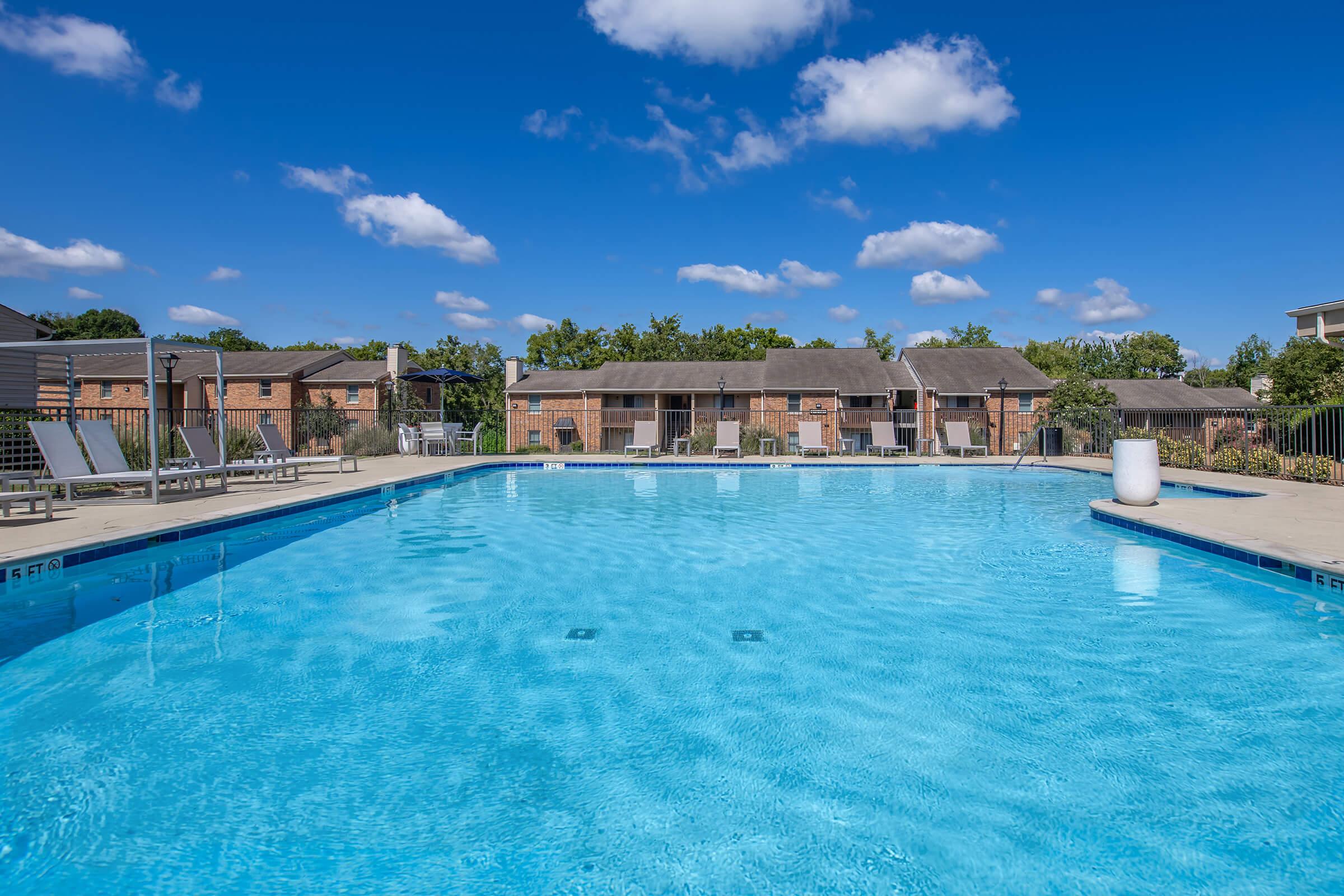 A clear blue swimming pool surrounded by lounging chairs under a bright blue sky with fluffy white clouds. The pool is bordered by brick buildings and greenery, creating a relaxing outdoor atmosphere.