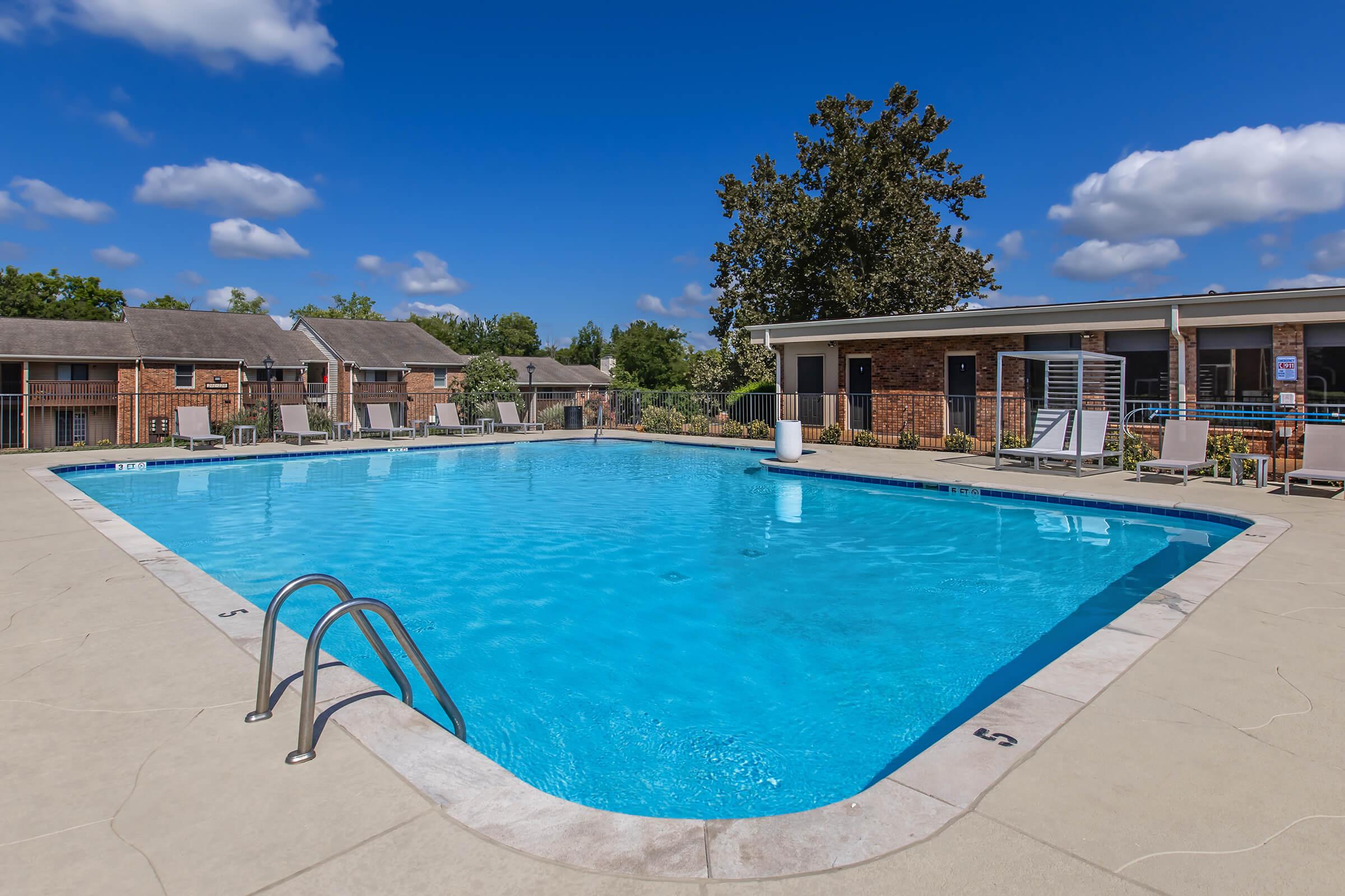 A clear blue swimming pool with steps leading into it, surrounded by concrete decking. In the background, there are several residential buildings and trees under a bright blue sky with scattered clouds. Loungers are positioned around the pool area for relaxation.