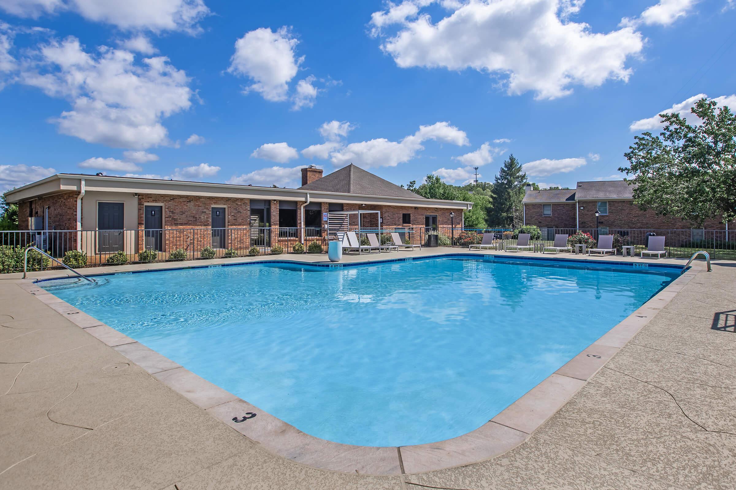 A sunny outdoor swimming pool surrounded by a concrete deck, featuring lounge chairs and a diving board. A brick building with multiple windows is visible in the background, along with trees and green shrubs. The sky is bright blue with fluffy white clouds.