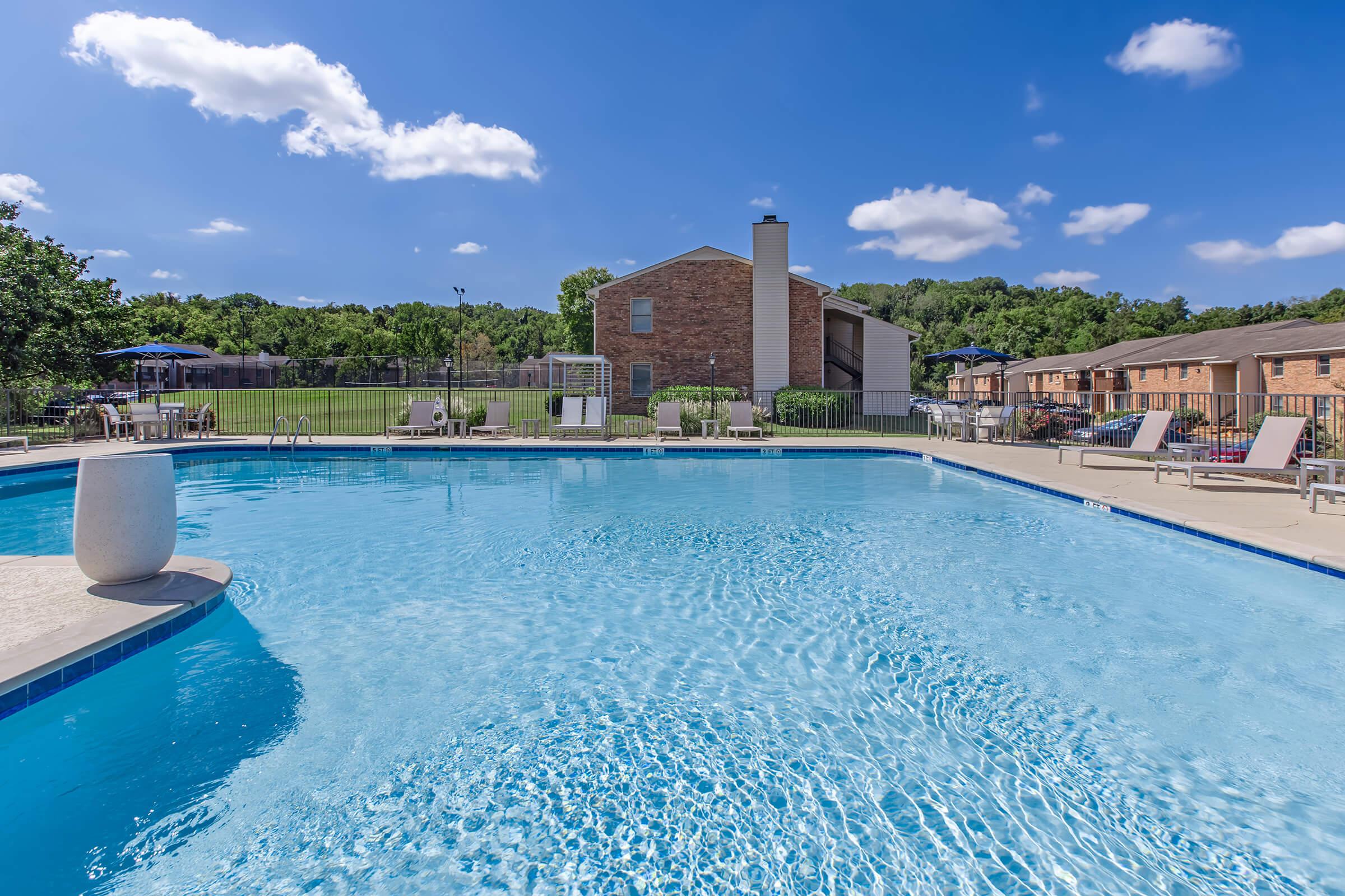 A clear blue swimming pool surrounded by lounge chairs and tables, with a brick building in the background. Lush green trees and a grassy area are visible beyond the pool, under a bright blue sky with a few fluffy white clouds.