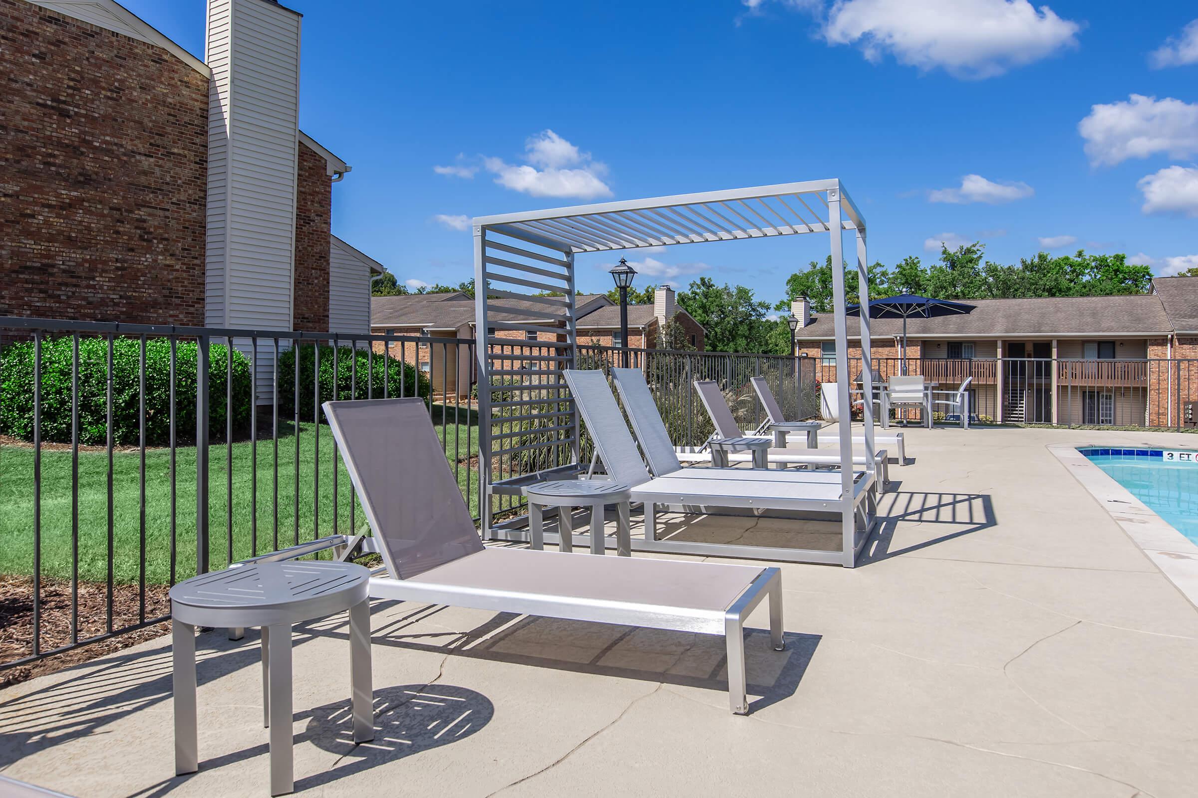 A sunny outdoor pool area featuring multiple lounge chairs and a shaded cabana. The pool is visible in the background, surrounded by a fence. Lush greenery and residential buildings are present, under a clear blue sky with a few clouds.