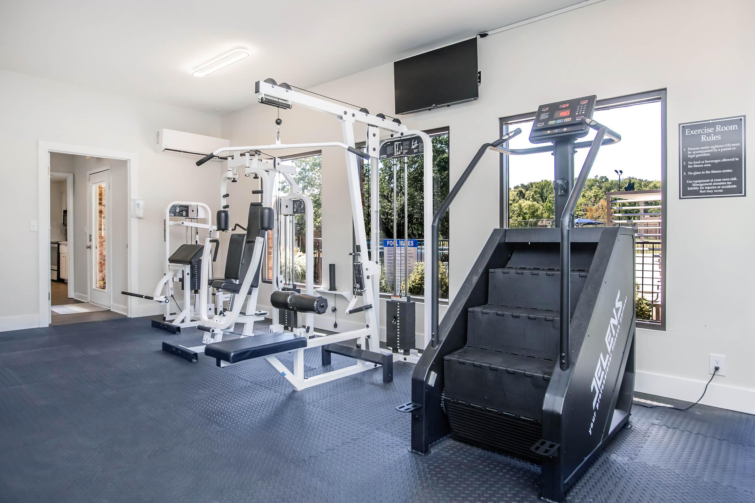 A modern gym space featuring a multi-station weight machine, a stair stepper, and a large window providing natural light. The room has a clean design with rubber flooring, a wall-mounted TV, and a sign detailing exercise room rules.