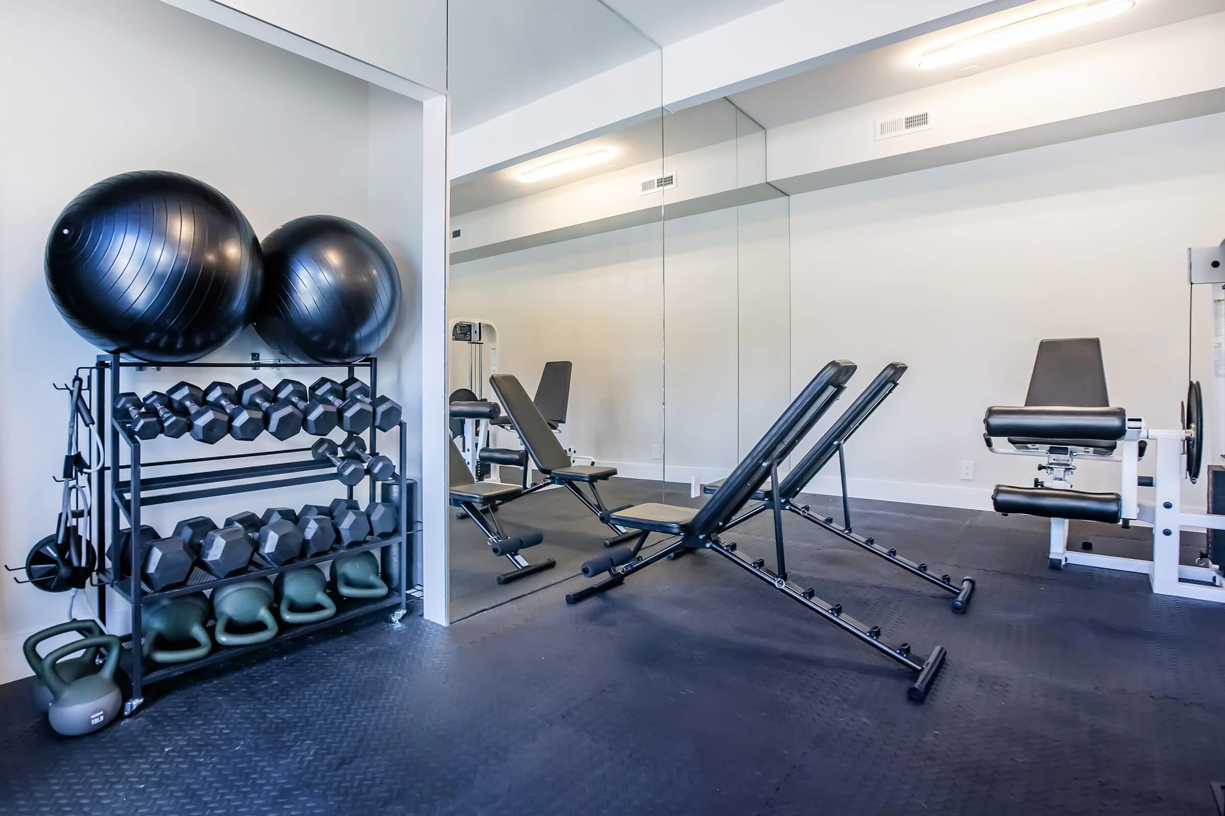 A modern gym interior featuring exercise equipment. On the left, a rack holds various dumbbells and kettlebells, while in the center, adjustable workout benches are positioned facing a mirror. The space is well-lit with sleek flooring, creating an inviting environment for fitness activities.