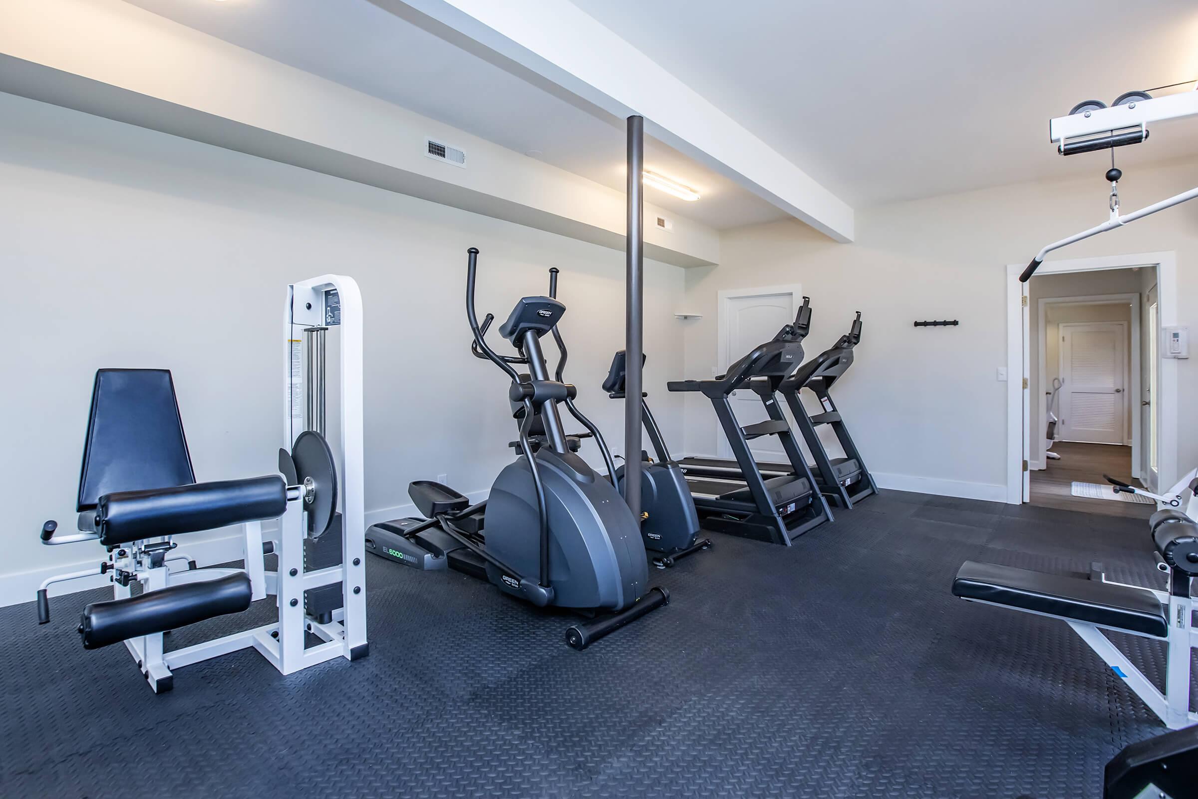 A modern fitness room featuring an elliptical machine, a leg press machine, and multiple treadmills. The space has rubber flooring, bright walls, and a mirror, creating a clean and inviting atmosphere for workouts. A door in the background leads to another area.