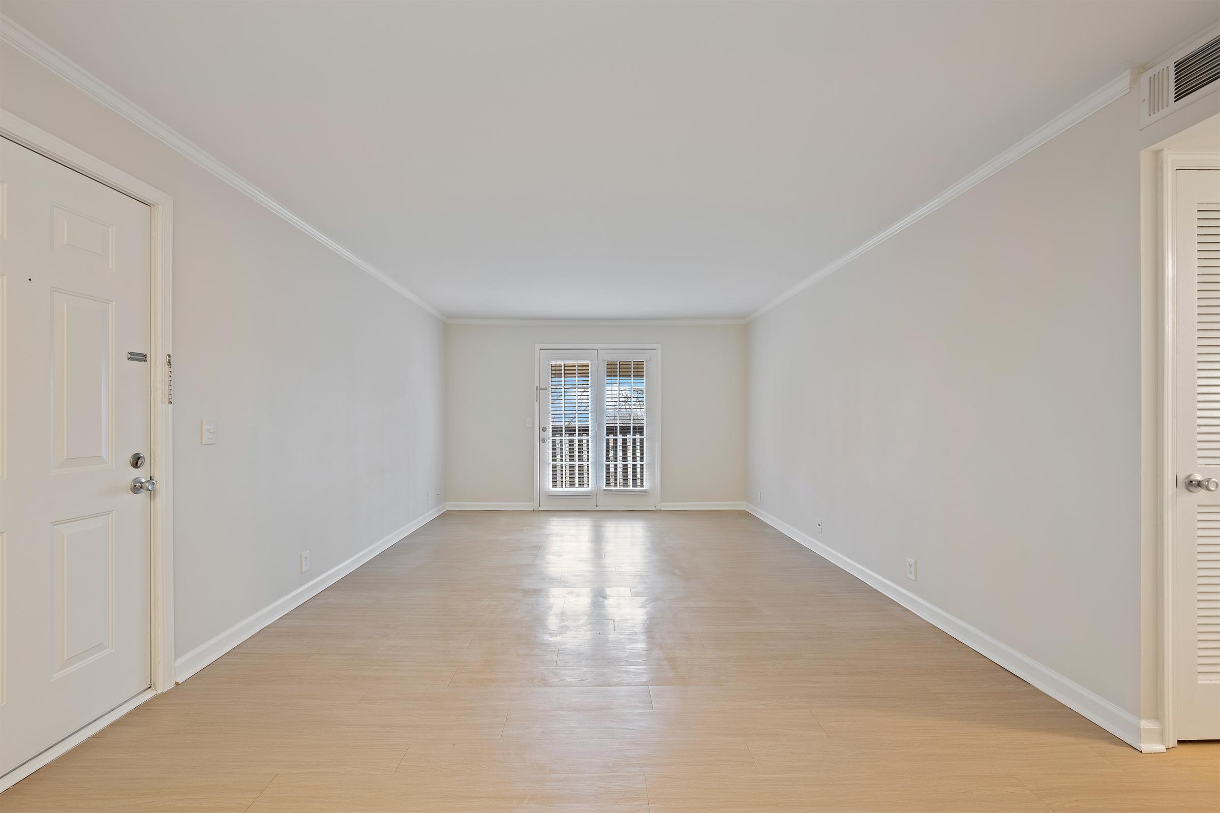 An empty, lightly-colored room with light wood flooring, featuring a door on the left and large windows with shutters at the far end. The walls are plain and neutral, giving a spacious and airy feel to the space.