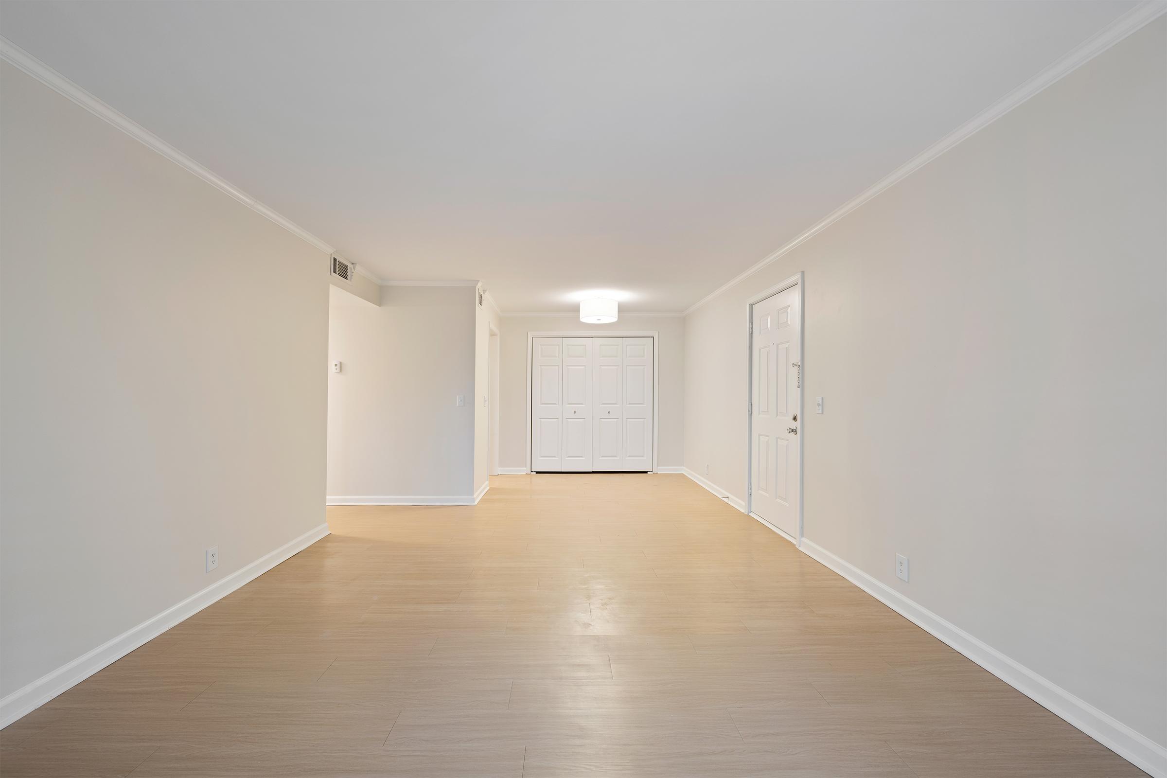 A spacious, empty room with light-colored walls and a pale wood floor. A modern light fixture hangs from the ceiling, and a set of white double doors is visible in the back, leading to a closet or another room. The overall atmosphere is bright and clean, ideal for decoration or furnishing.
