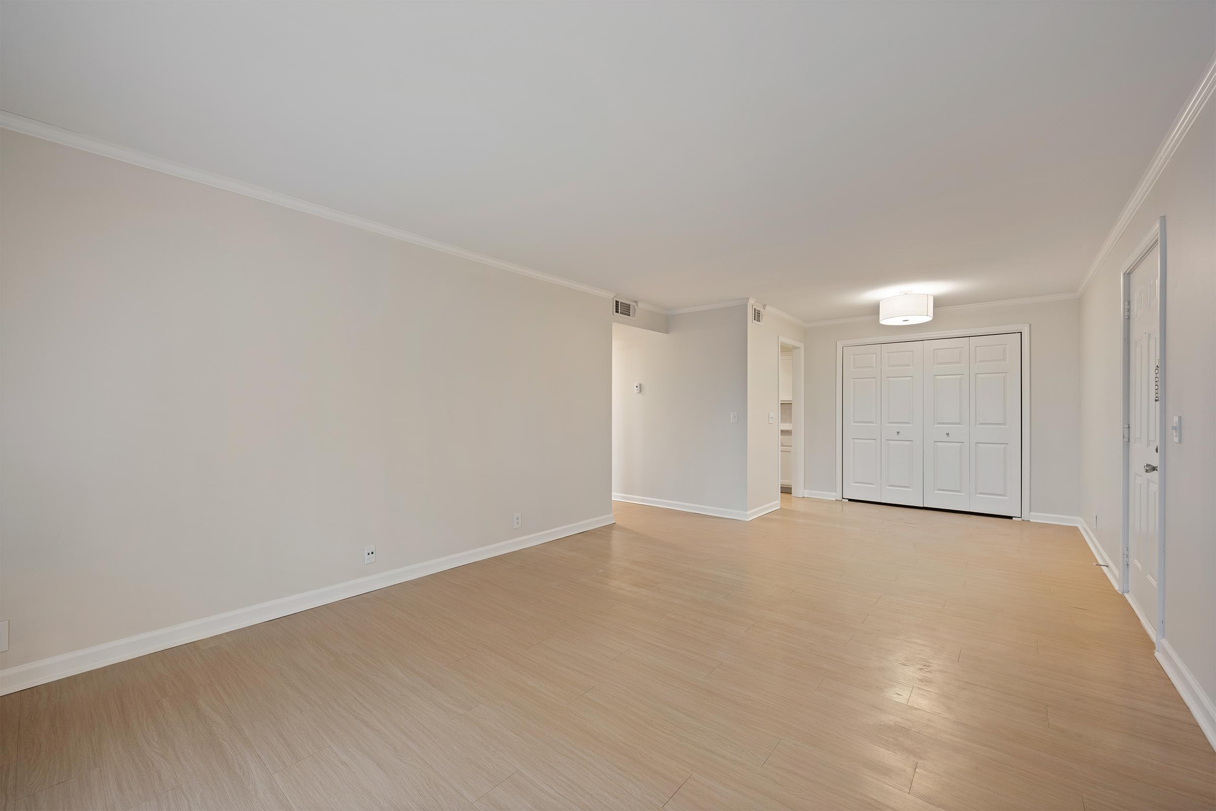A spacious, empty room with light-colored walls and smooth beige flooring. A doorway leads to another room that appears to have built-in closets. Natural light brightens the area, and the ceiling features a modern light fixture. The space is minimalistic, offering potential for various interior design possibilities.