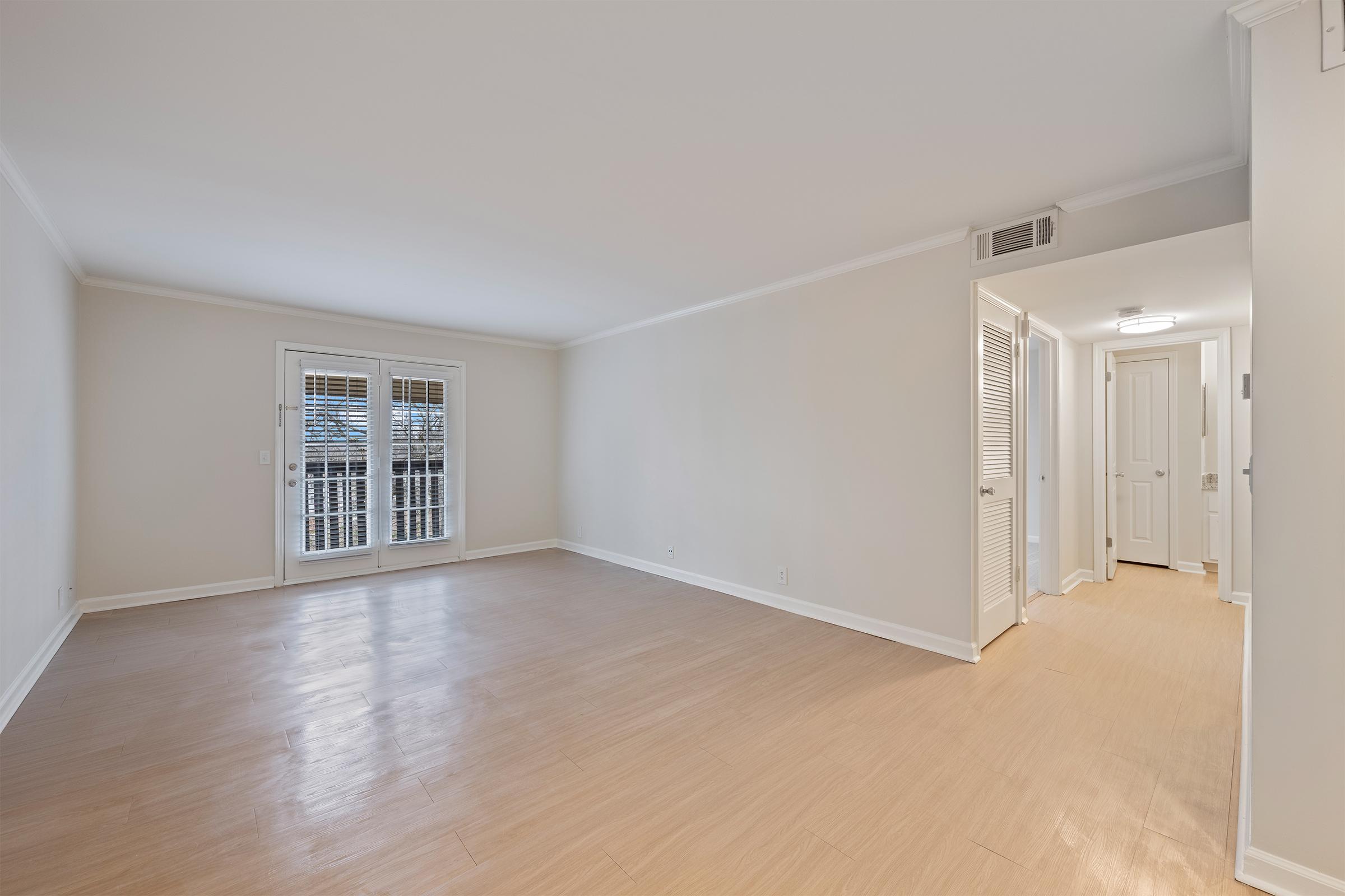 A bright, empty room with light wood flooring and white walls. There are large windows with white shutters on one side, allowing natural light in. A hallway is visible on the right, leading to closed doors, suggesting additional spaces in the apartment.
