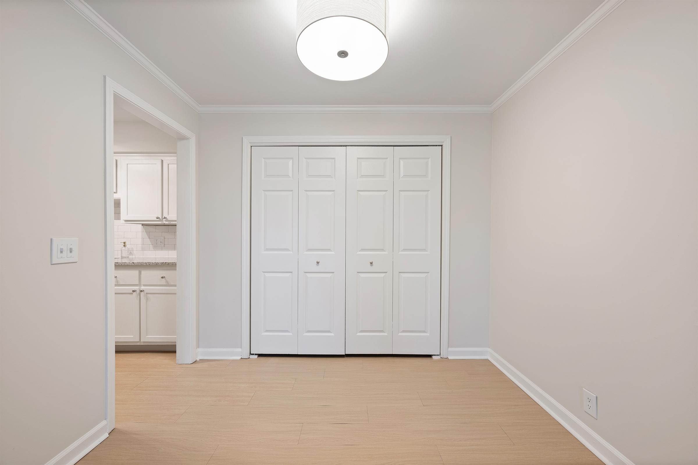A well-lit, neutral-toned room featuring a light fixture on the ceiling. In the foreground, there are double doors leading to a closet, while an open entryway on the left provides access to a kitchen area visible in the background. The flooring is light wood, creating a warm and inviting atmosphere.