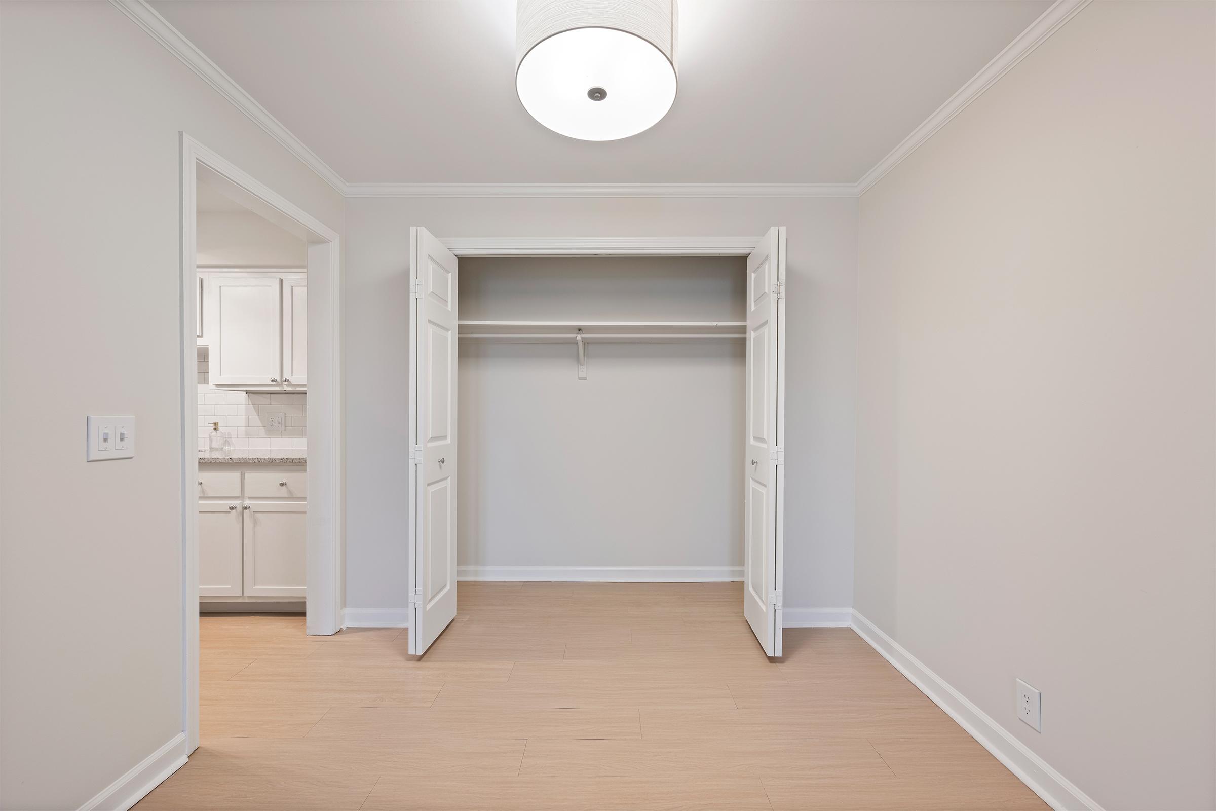 A well-lit, empty room featuring an open closet with white doors. The walls are painted light gray, and there is a modern light fixture on the ceiling. To the left, an entryway leads into a kitchenette with white cabinetry. The flooring is a light wood tone, creating a clean and spacious atmosphere.