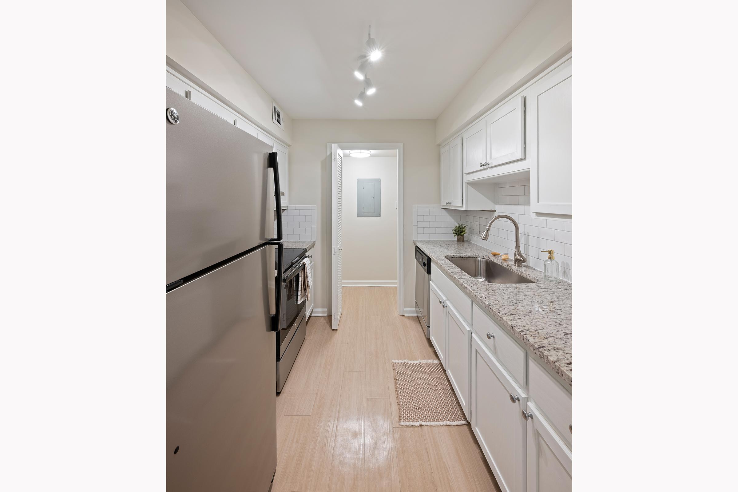 A modern kitchen featuring stainless steel appliances, including a refrigerator and oven. The countertops are made of granite, and there are white cabinets with a tiled backsplash. Natural light fills the space, and there is a small rug on the floor. The entrance to another room is visible at the end of the hallway.