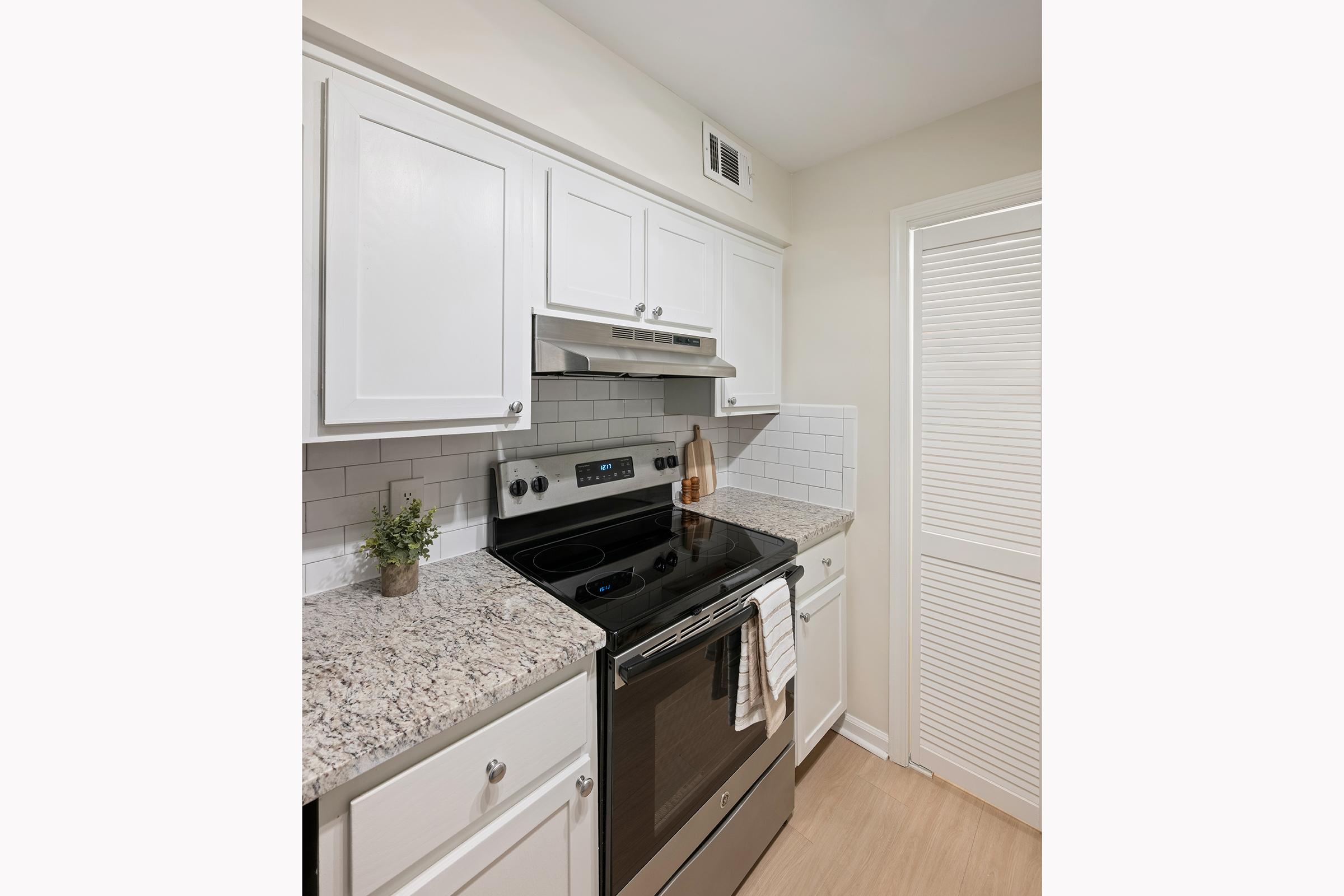 A modern kitchen featuring white cabinetry, a black stove with a stainless steel hood, and granite countertops. A small potted plant sits on the counter, and the space includes a white louvered door. The overall design is clean and contemporary with a bright, neutral color scheme.