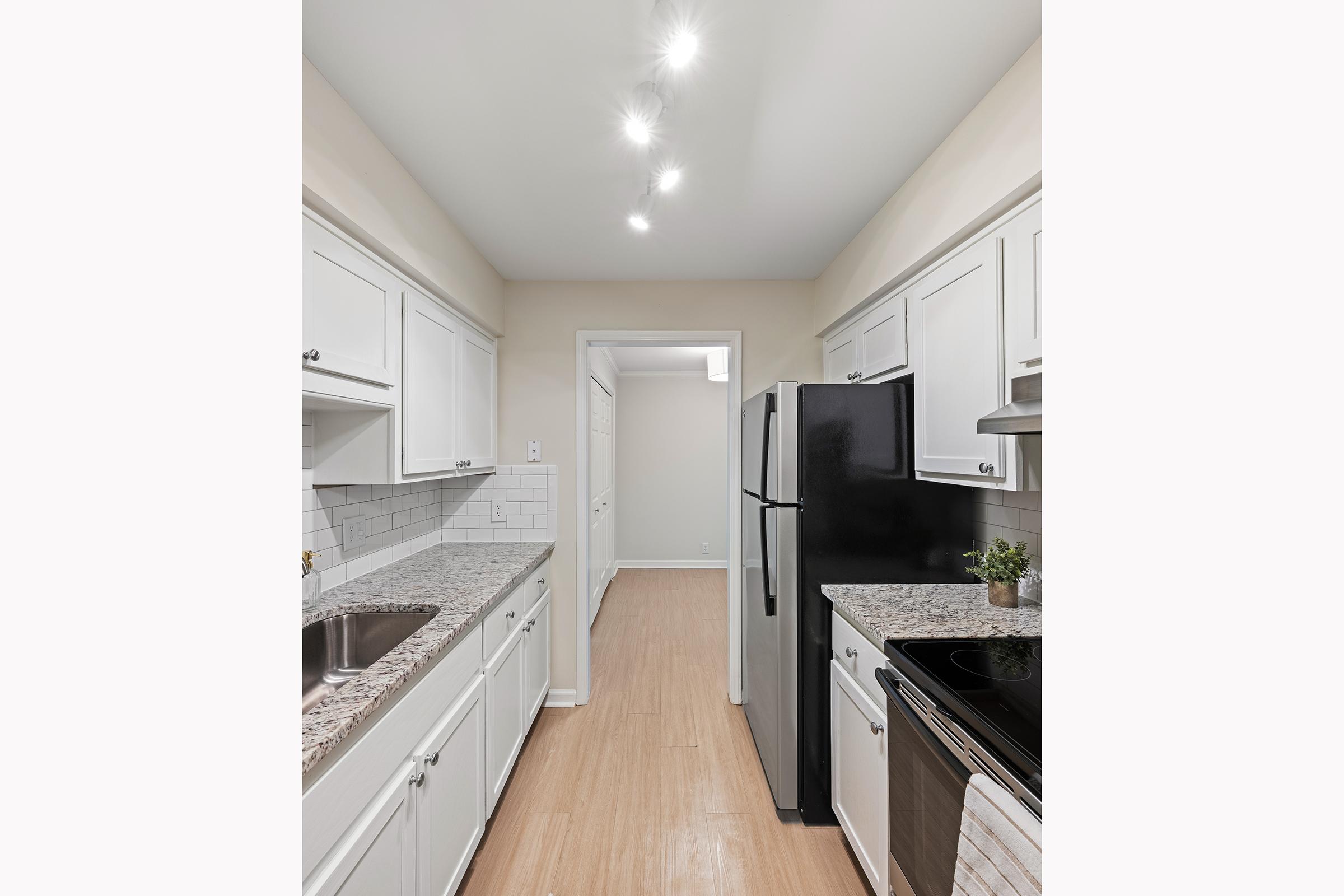 A bright, modern kitchen featuring white cabinetry, a stainless steel oven, and a black refrigerator. The countertops are made of speckled granite, and the space has recessed lighting. The flooring is light-colored, enhancing the airy feel of the room. A small plant adds a touch of greenery.