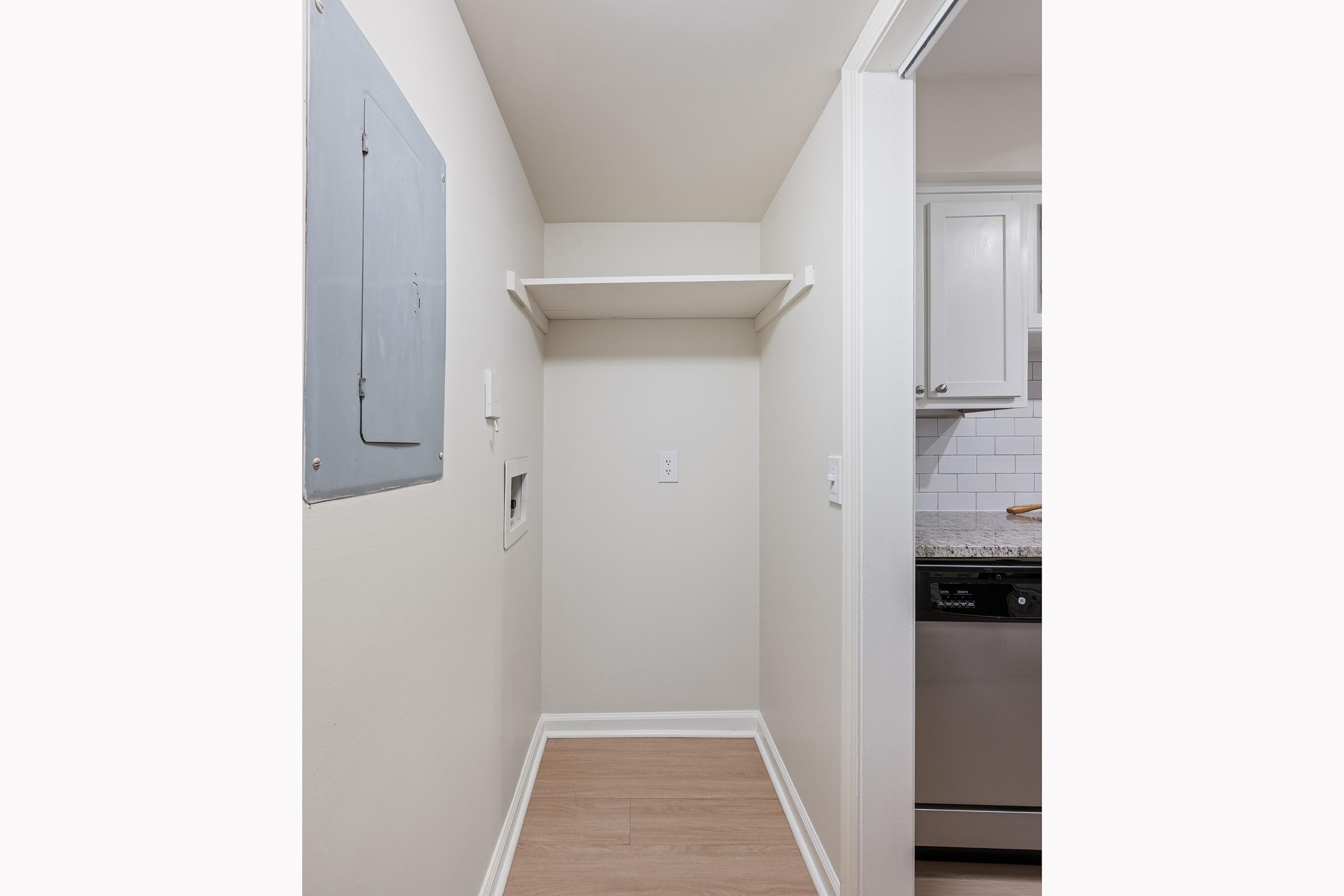 A small, empty utility space with light-colored walls and a shelf on one side. The floor is a light wood laminate, and an electrical panel is visible on the left wall. In the background, there is a glimpse of a kitchen area with gray cabinets and white tile.