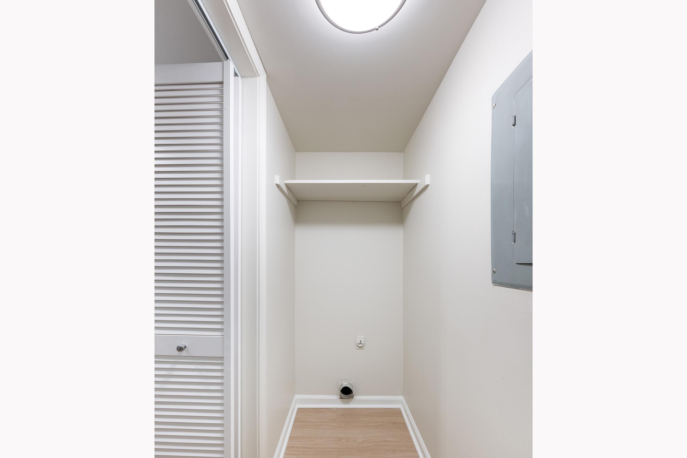A small, empty closet space featuring a light fixture on the ceiling, a single shelf mounted on the wall, and a vent or outlet on the floor. The adjacent wall has a panel or door with horizontal slats, and the flooring is light-colored wood.