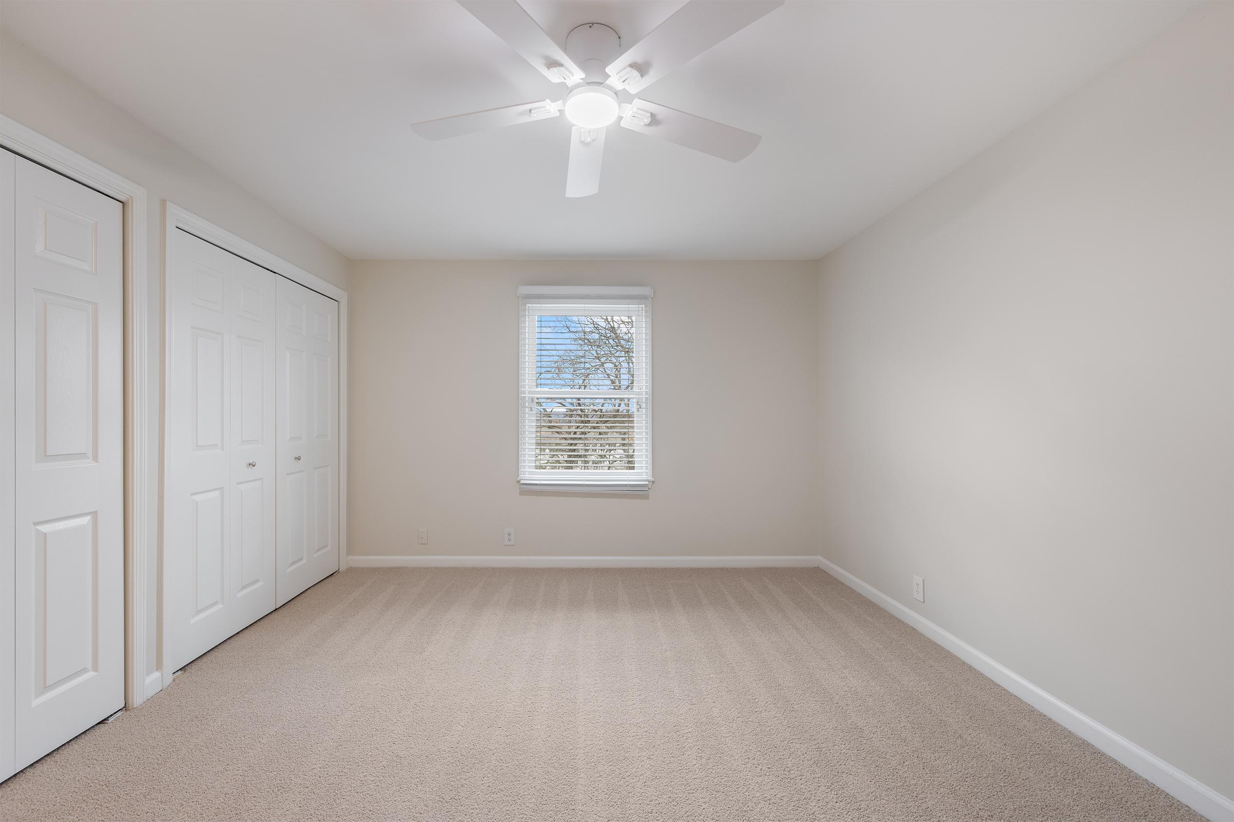 A bright, empty room with light beige walls and carpet. It features a ceiling fan, a window with a view of trees outside, and two white closet doors. The room has a simple, clean aesthetic, making it suitable for various uses.