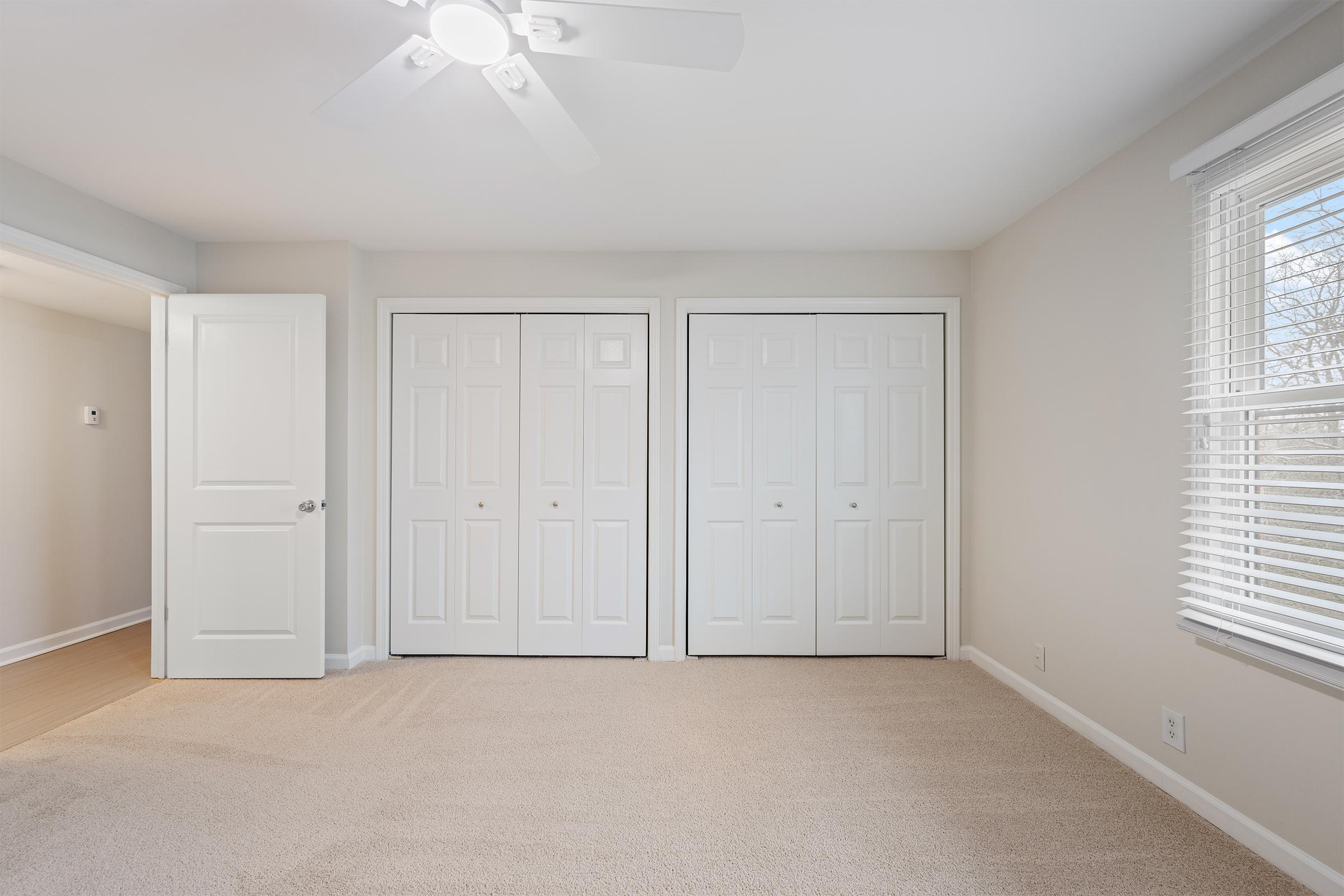 A spacious room with light-colored walls and carpeted flooring. It features two sets of closed double doors for closets, a ceiling fan on the white ceiling, and a window with blinds allowing natural light to enter the space. The room appears tidy and well-lit, ideal for various uses.