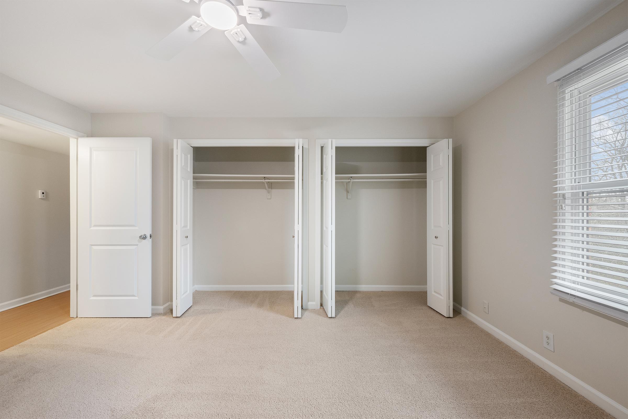 A well-lit, empty room featuring two open closet doors on one side, beige carpeted flooring, and light-colored walls. A ceiling fan is visible. Natural light filters in through a window, creating a bright and airy atmosphere. The space is ready for furniture or decoration.