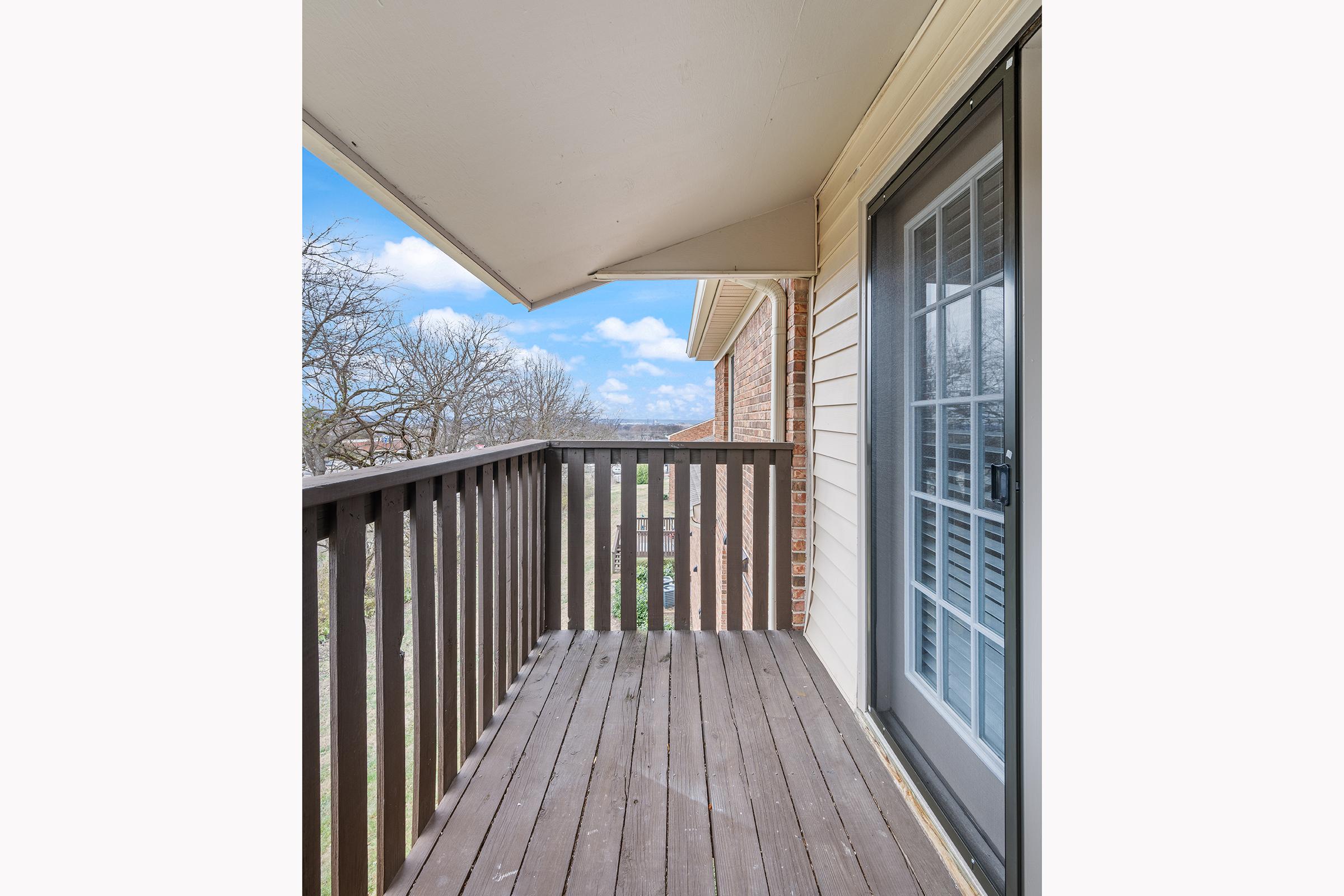 A balcony with wooden flooring and a railing, featuring a door leading into an interior space. The view beyond includes trees and a clear blue sky, creating a serene atmosphere. The building is constructed from light-colored materials, and the ceiling is a light shade, enhancing the open feel of the area.