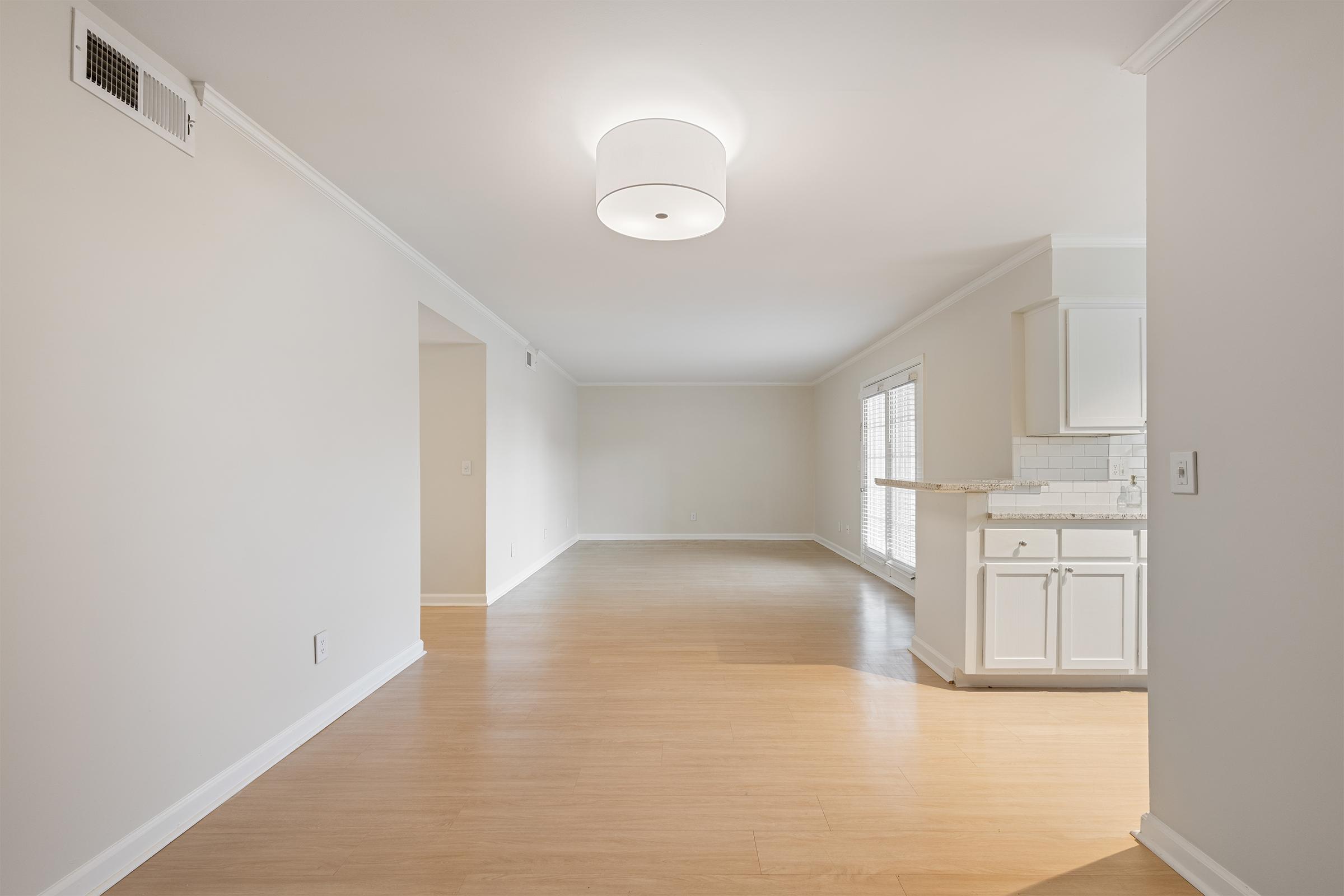 A spacious, well-lit room featuring a light-colored, empty interior with wooden flooring. On the right, there is a small kitchen area with white cabinets and a countertop. Large windows allow natural light to enter, while a modern ceiling light fixture illuminates the space.
