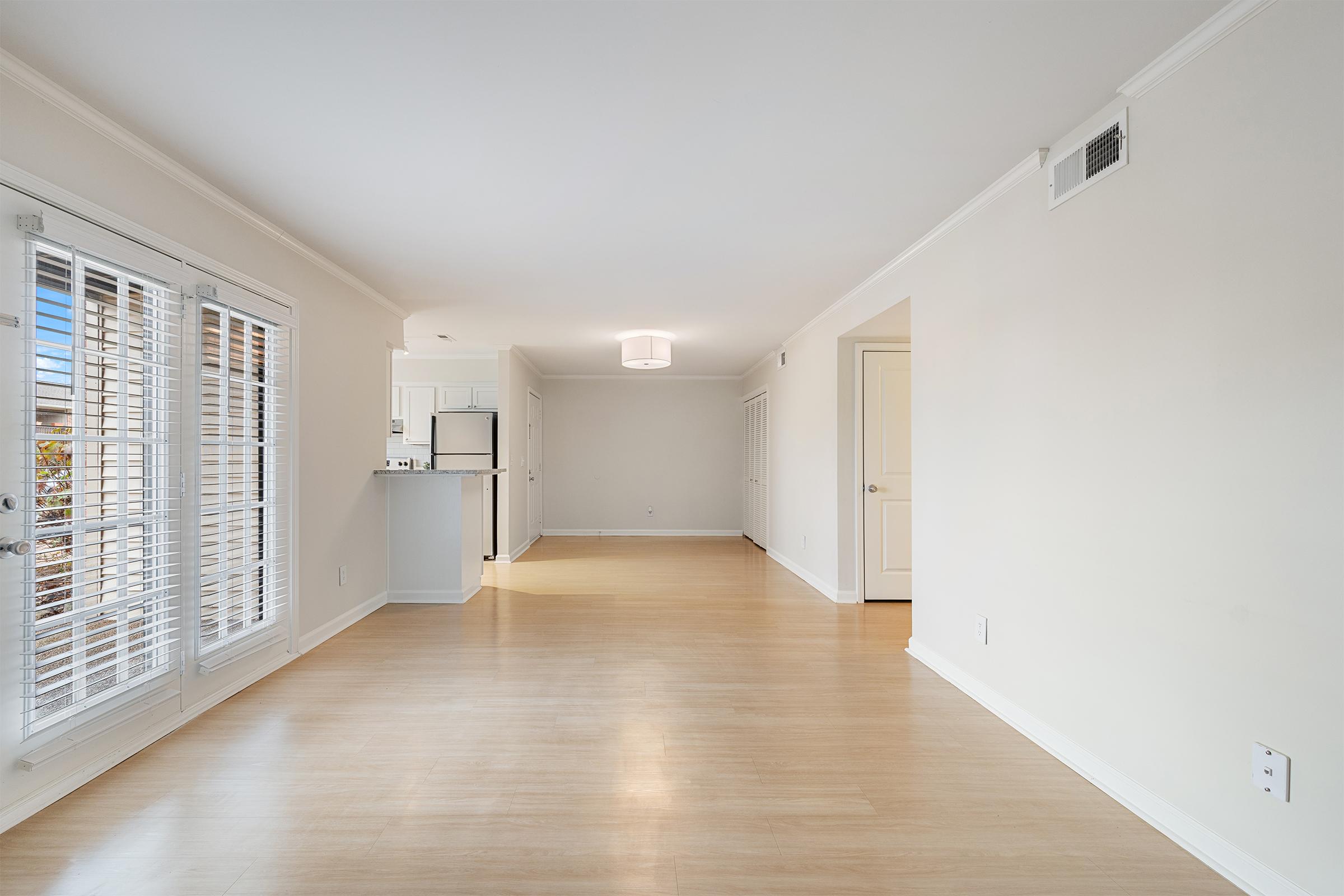 Spacious, empty room with light-colored hardwood flooring, featuring large windows with white blinds. There is a modern light fixture on the ceiling, and an open doorway leads to a kitchen area in the background. The walls are painted a soft neutral color, creating a bright and airy atmosphere.