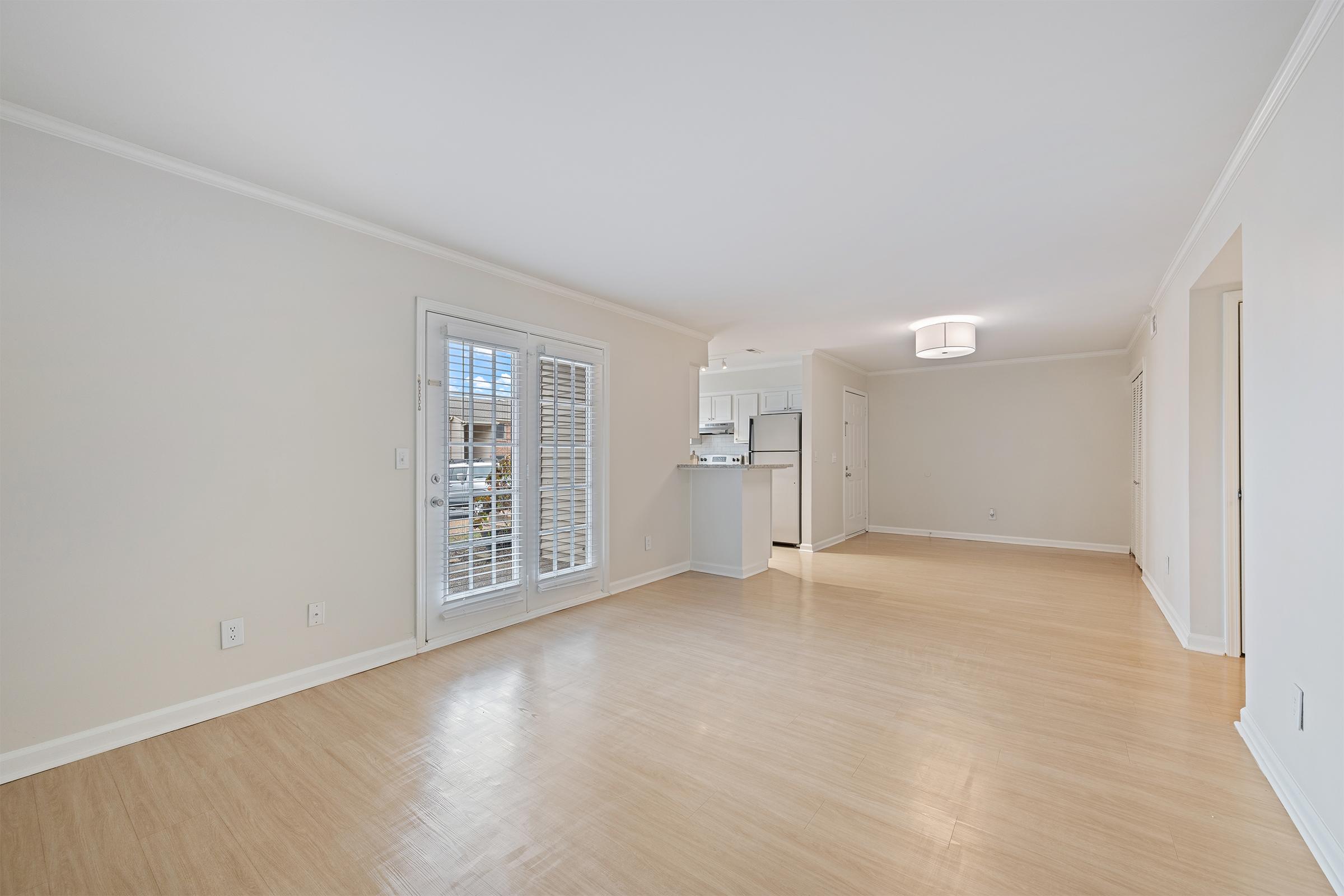 Bright and airy room with light-colored walls and hardwood flooring. There are white plantation shutters on two windows and a modern light fixture on the ceiling. A doorway leads to a kitchen area visible in the background, complete with white cabinetry. The space is empty and inviting, perfect for various furnishings.