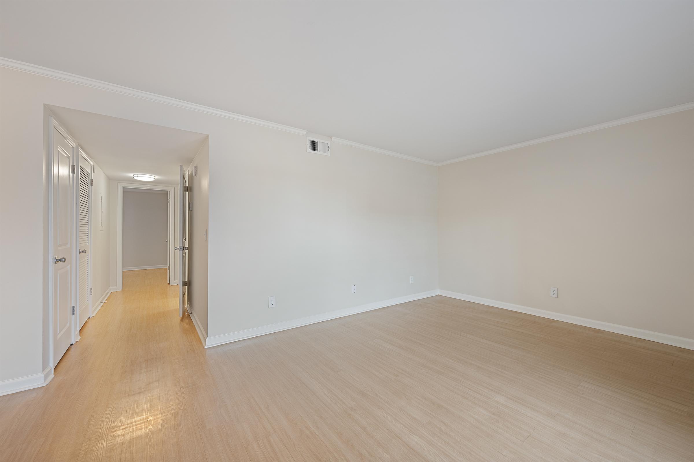 A bright, empty room with light-colored walls and bamboo flooring. To the left, there's a hallway leading to other rooms with doors, and the space is well-lit by natural light. The overall atmosphere is clean and spacious, perfect for decoration or furnishing.
