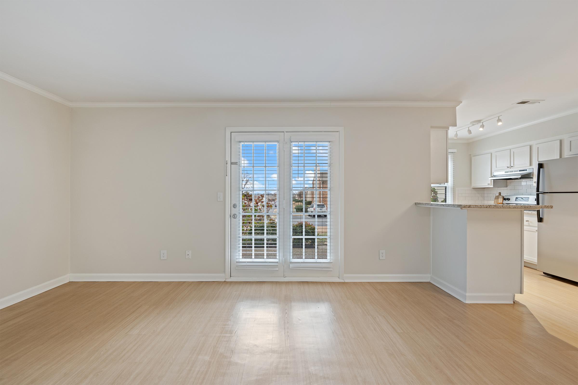 A bright, spacious living area featuring light-colored walls and hardwood flooring. It has large sliding glass doors with white shutters that lead outside, and a kitchen area visible in the background with modern appliances. The overall atmosphere is clean and welcoming, ideal for relaxation and entertaining.