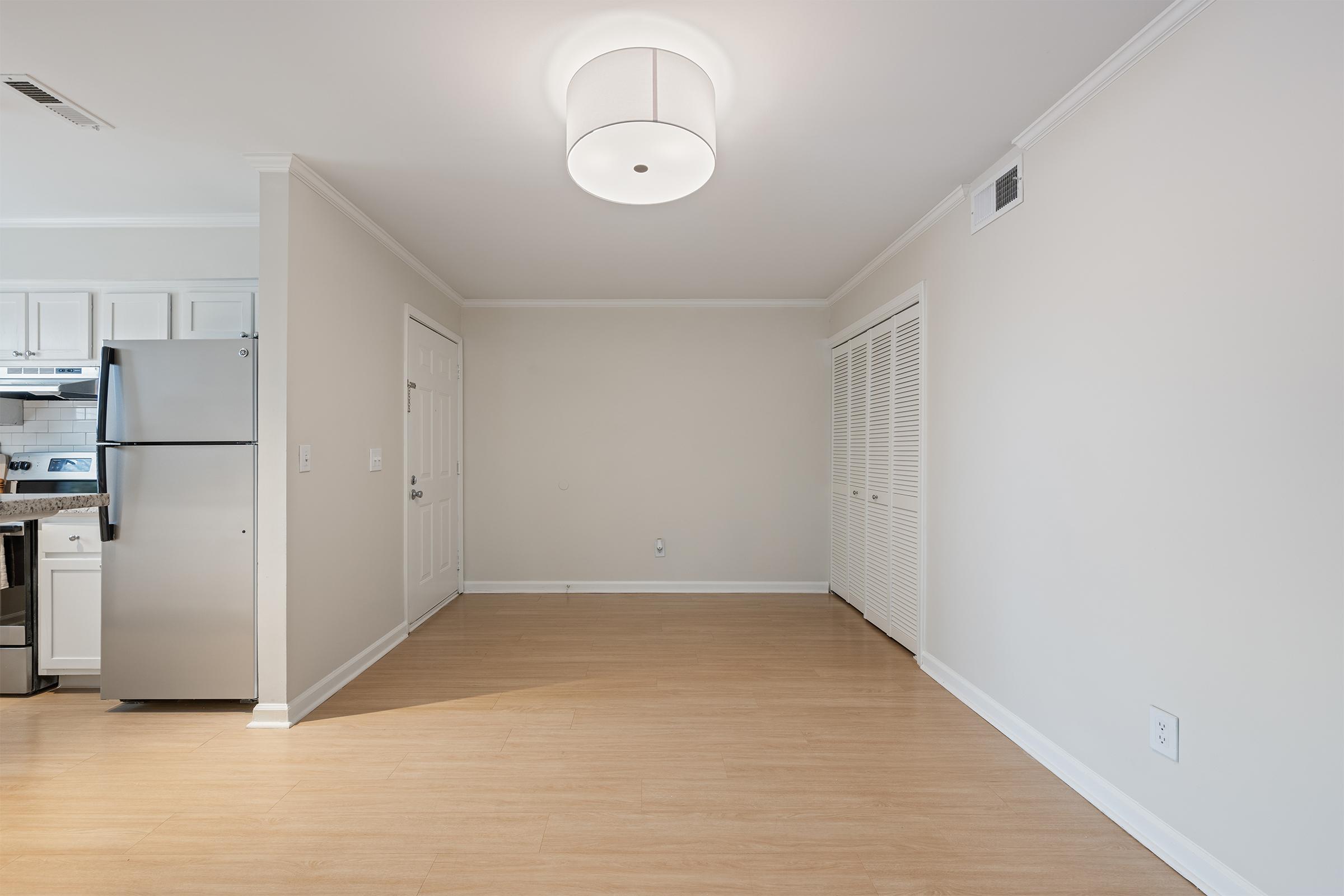 Interior of an empty room featuring light-colored walls and a wooden floor. A modern light fixture is visible on the ceiling. To the left, there is a kitchen with stainless steel appliances. The room has a door leading to another space and a closet with sliding doors. Natural light brightens the area.