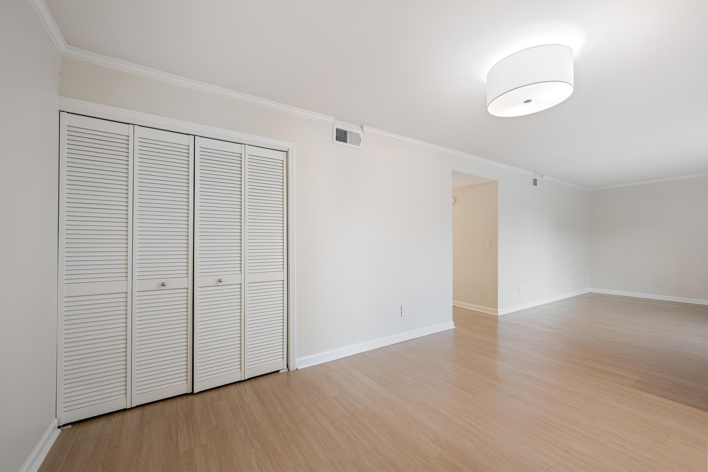 A bright, empty room featuring light-colored walls and polished wooden flooring. A large, modern ceiling light fixture is visible, along with a white closet with louvered doors on the left side. The room has an open layout, leading to another area in the background. Natural light enhances the spacious feel.