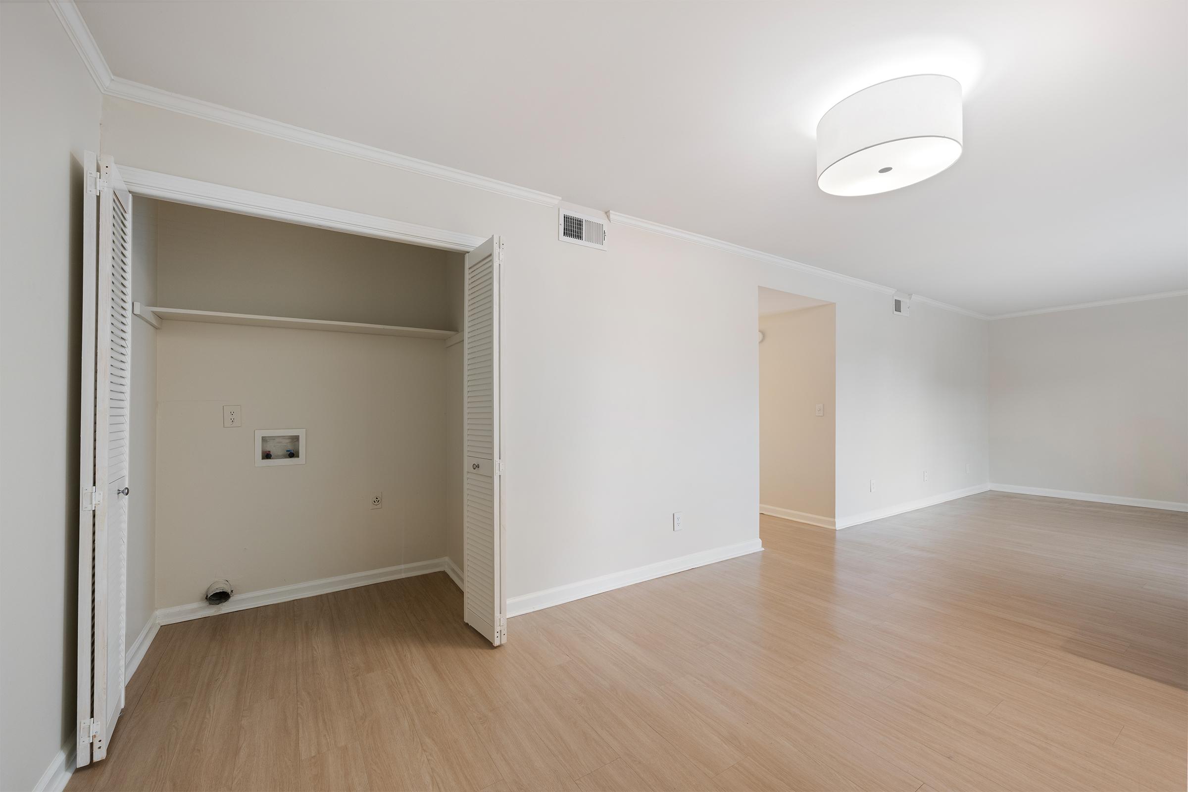 A spacious, empty room with light-colored walls and wooden flooring. There is an illuminated ceiling fixture and a small closet with double doors on the left. The room has a minimalist design, offering ample natural light and a clear view of an adjoining room.