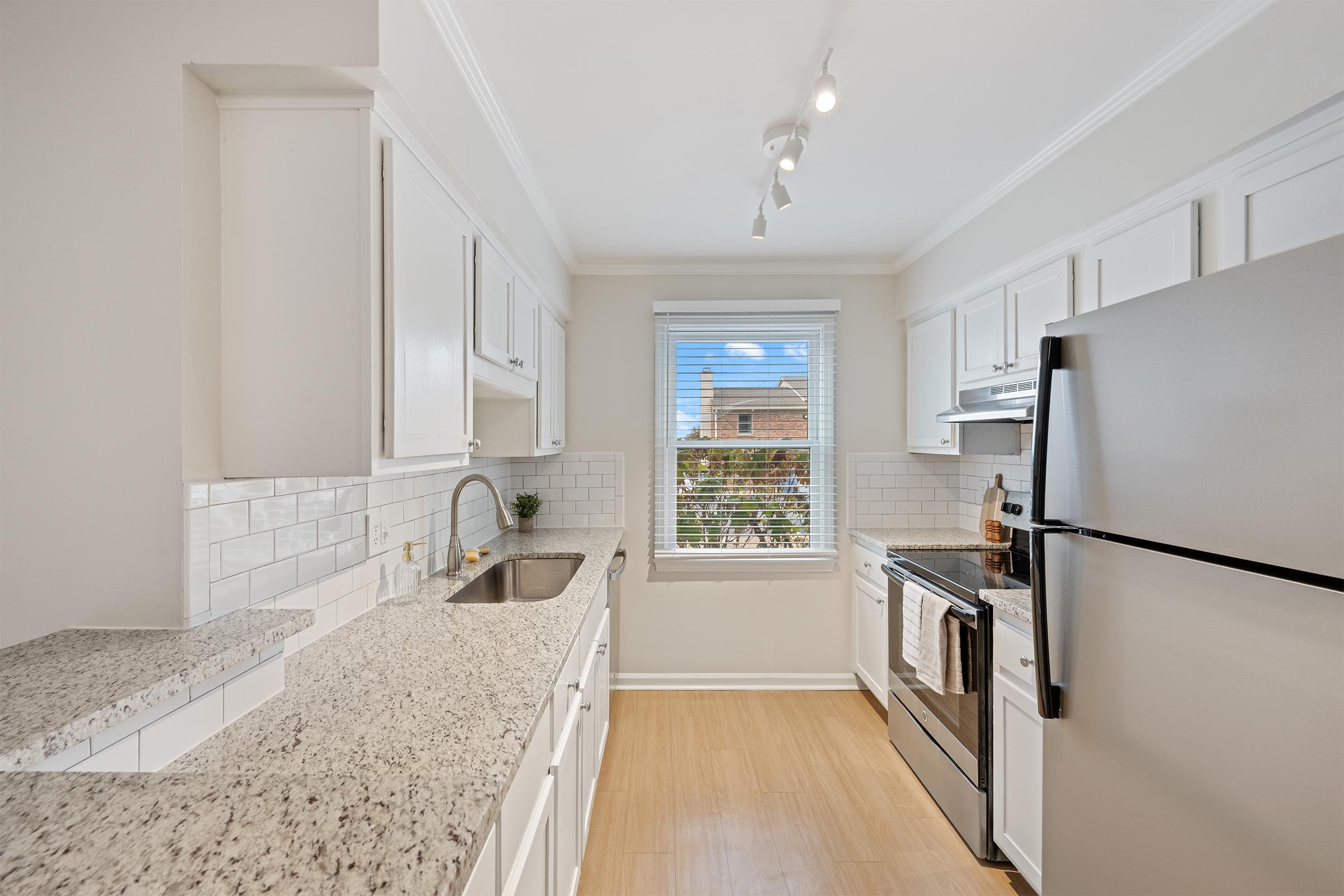 A bright and modern kitchen featuring white cabinets, a stainless steel refrigerator, and a stove. The countertop is made of light granite, and there's a window with a view outside. The kitchen has natural light and a clean, minimalist design with a cozy ambiance.