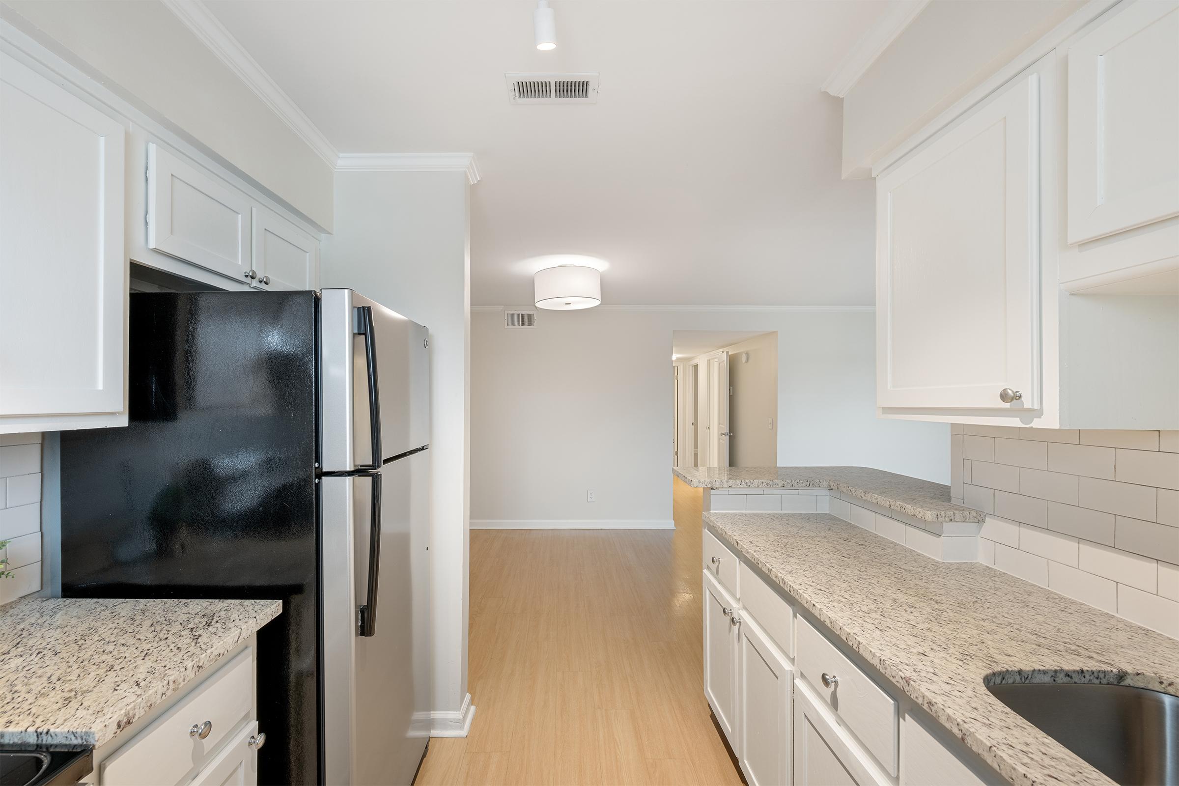 Modern kitchen featuring white cabinetry, black refrigerator, and stainless steel appliances. It has a granite countertop and a sink, with ample natural light. The open layout connects to a hallway leading to other rooms, showcasing a neutral color scheme and clean design.