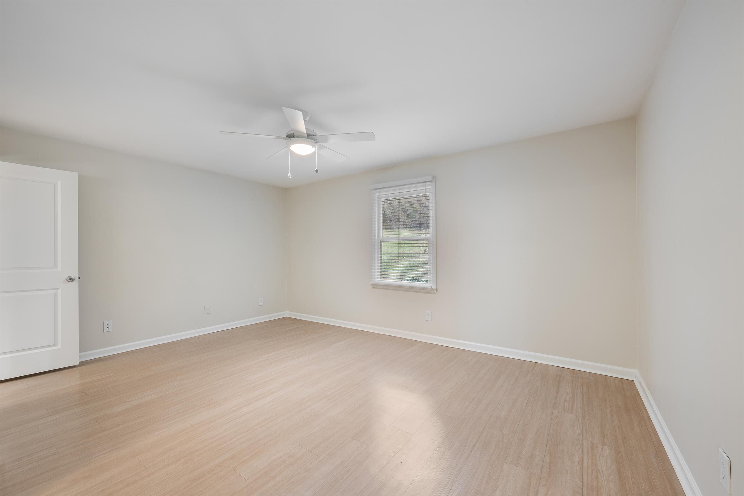 A spacious, empty room with light-colored walls and a ceiling fan. There is a single window allowing natural light, and the floor is made of wood or laminate. A white door is partially open, suggesting access to another area. The room appears clean and well-maintained.