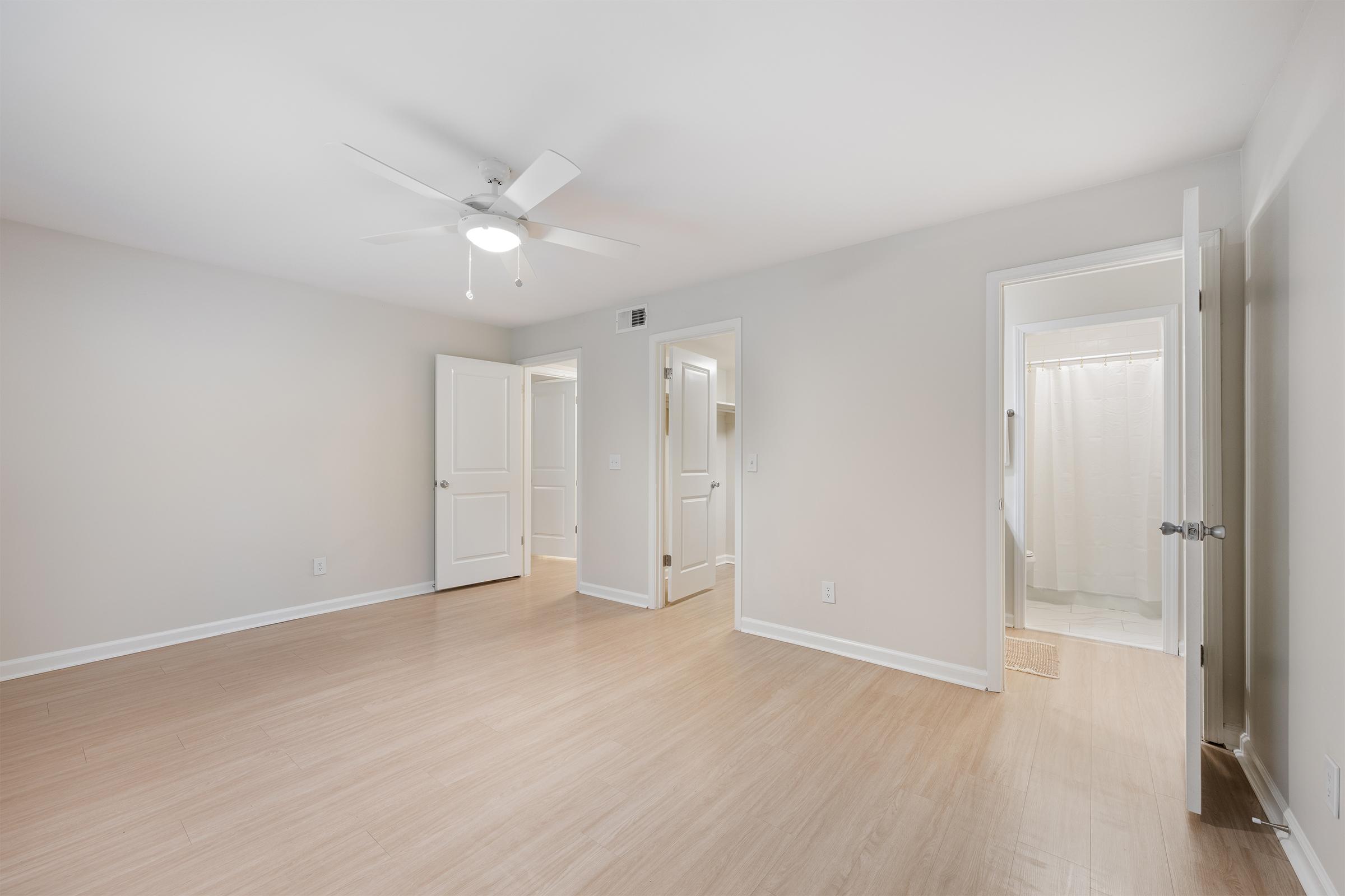 A spacious, well-lit room with light wooden flooring and pale gray walls. It features a ceiling fan and white doors leading to a bathroom and another room. Natural light enters through the doorway, creating a bright and inviting atmosphere.