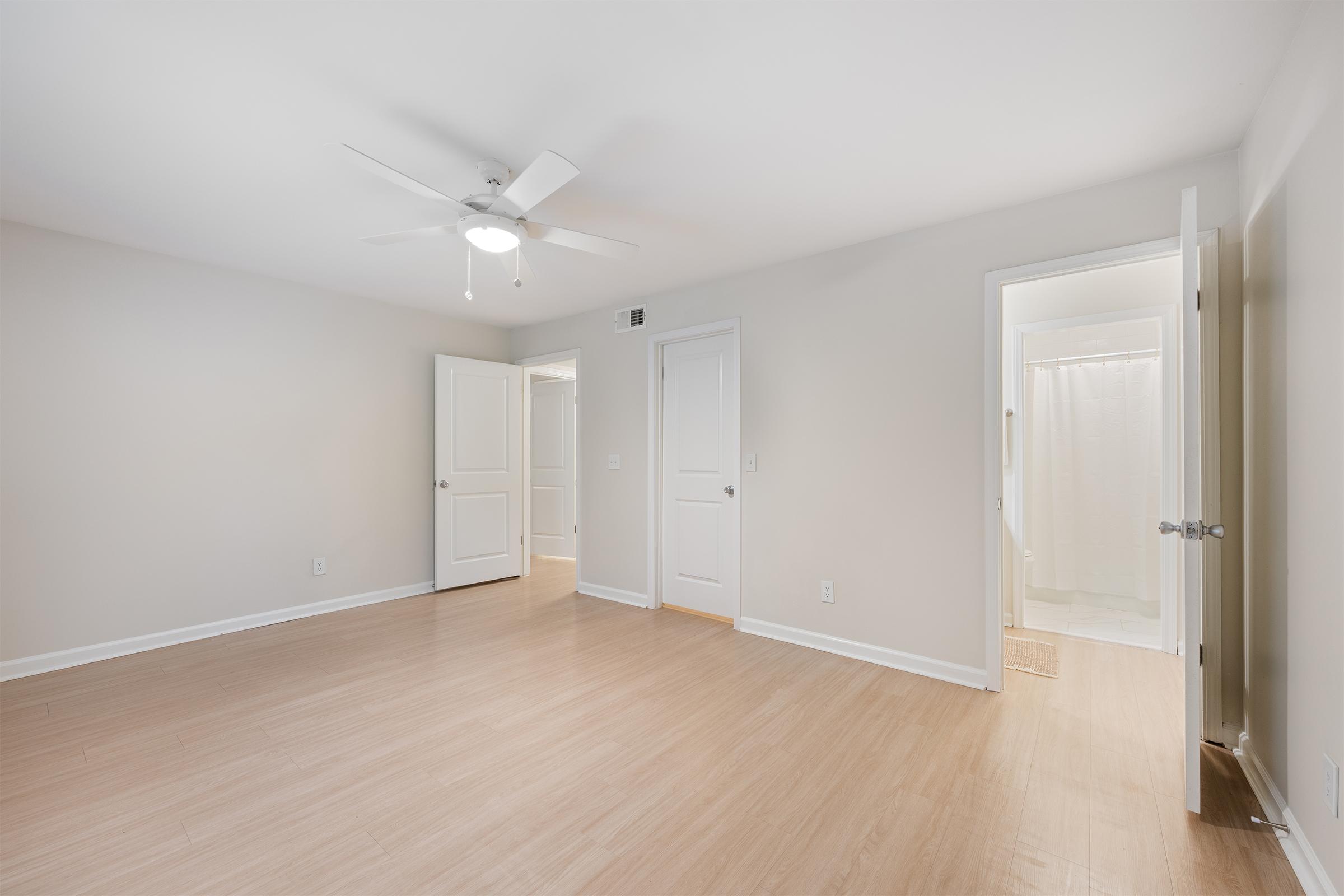 A spacious and empty room with light-colored walls and a ceiling fan. Two doors lead to adjacent rooms, and one door opens to a bathroom with a shower. The floor is light wood, providing a clean and open atmosphere. Natural light is visible, enhancing the bright and airy feeling of the space.