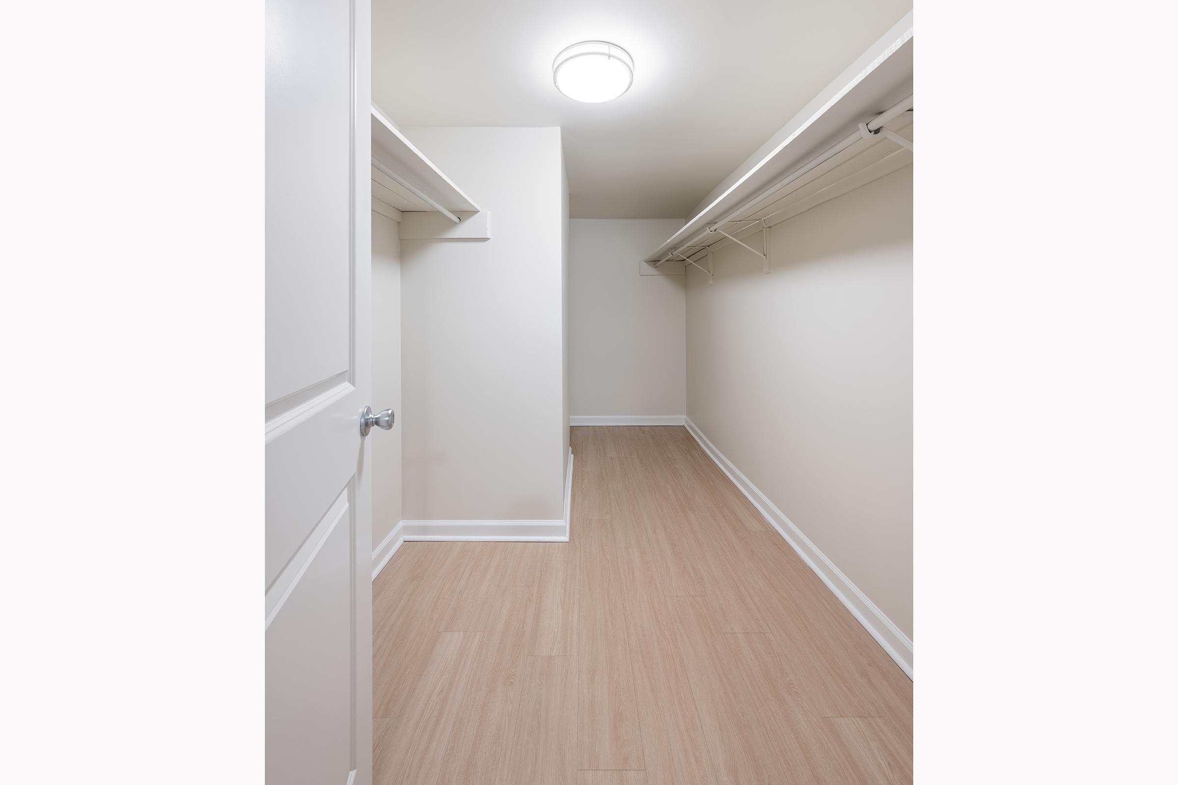 A spacious, empty walk-in closet with light-colored walls and wood-like flooring. The closet features two hanging racks on either side and a ceiling light fixture, providing natural brightness to the space. Ideal for storage or organization.