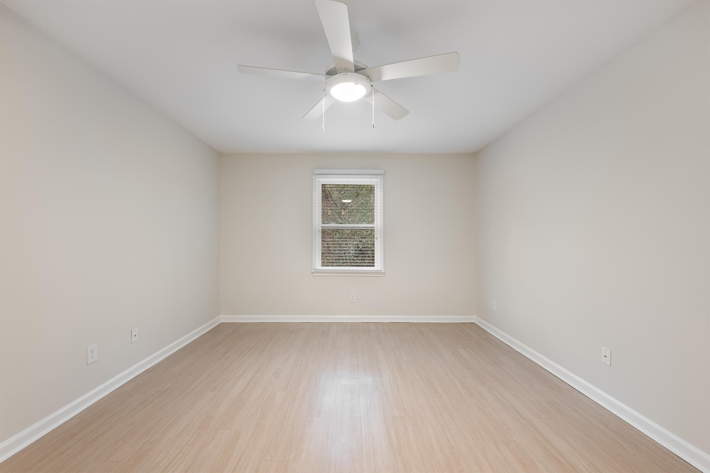 Empty room with light beige walls and a ceiling fan. A single window provides natural light, and the floor is made of light-colored laminate. The space is uncluttered and ready for furnishing.