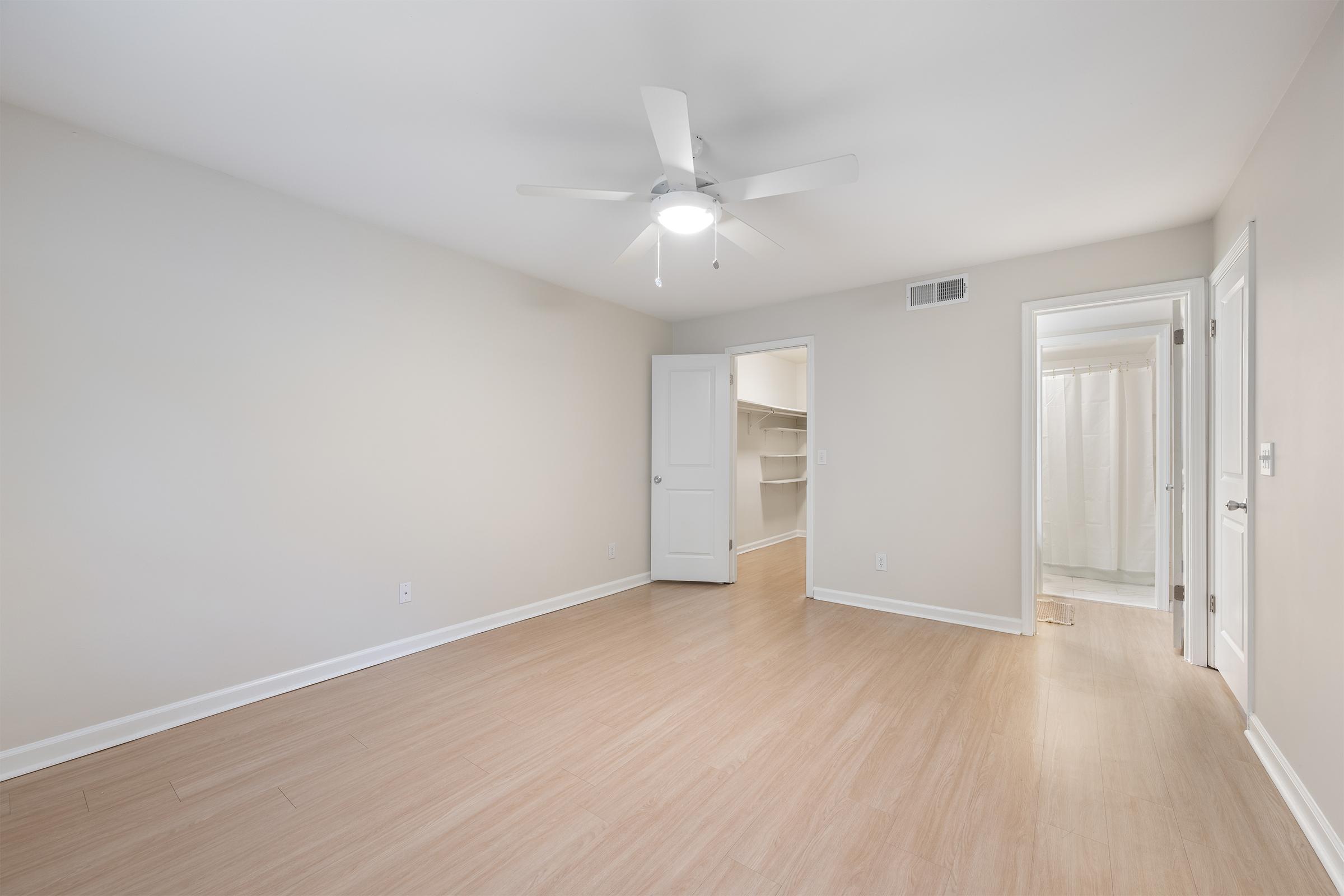 Empty room with light-colored walls and hardwood flooring. A ceiling fan is mounted in the center, and there is a closet on one side. A doorway leads to a bathroom area, and natural light enters the space, creating a bright and open atmosphere.