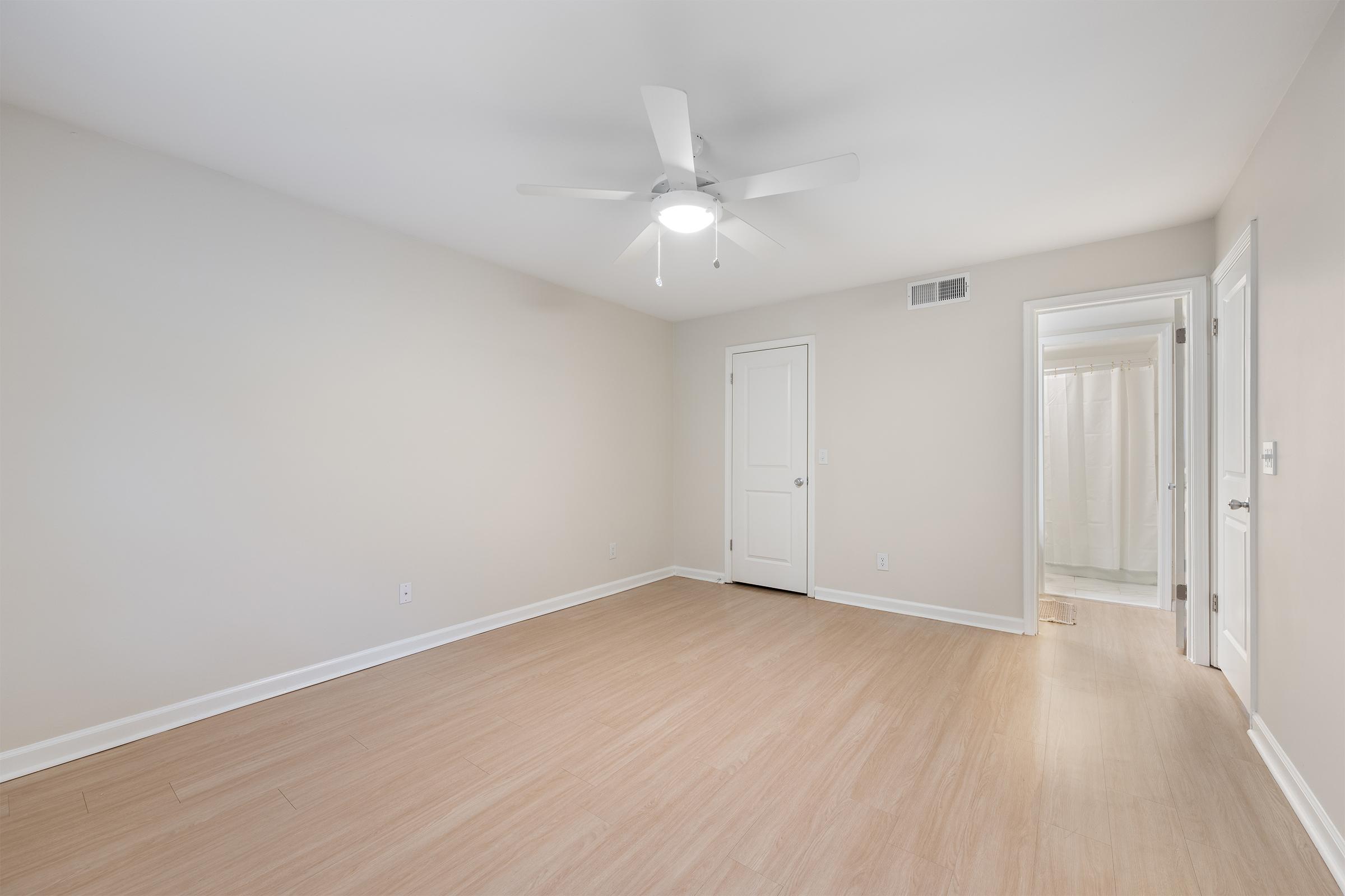 Empty bedroom featuring light-colored walls and a ceiling fan. The floor is made of light wood laminate. There is a door leading to a bathroom in the background, and the room has a bright, airy feel.