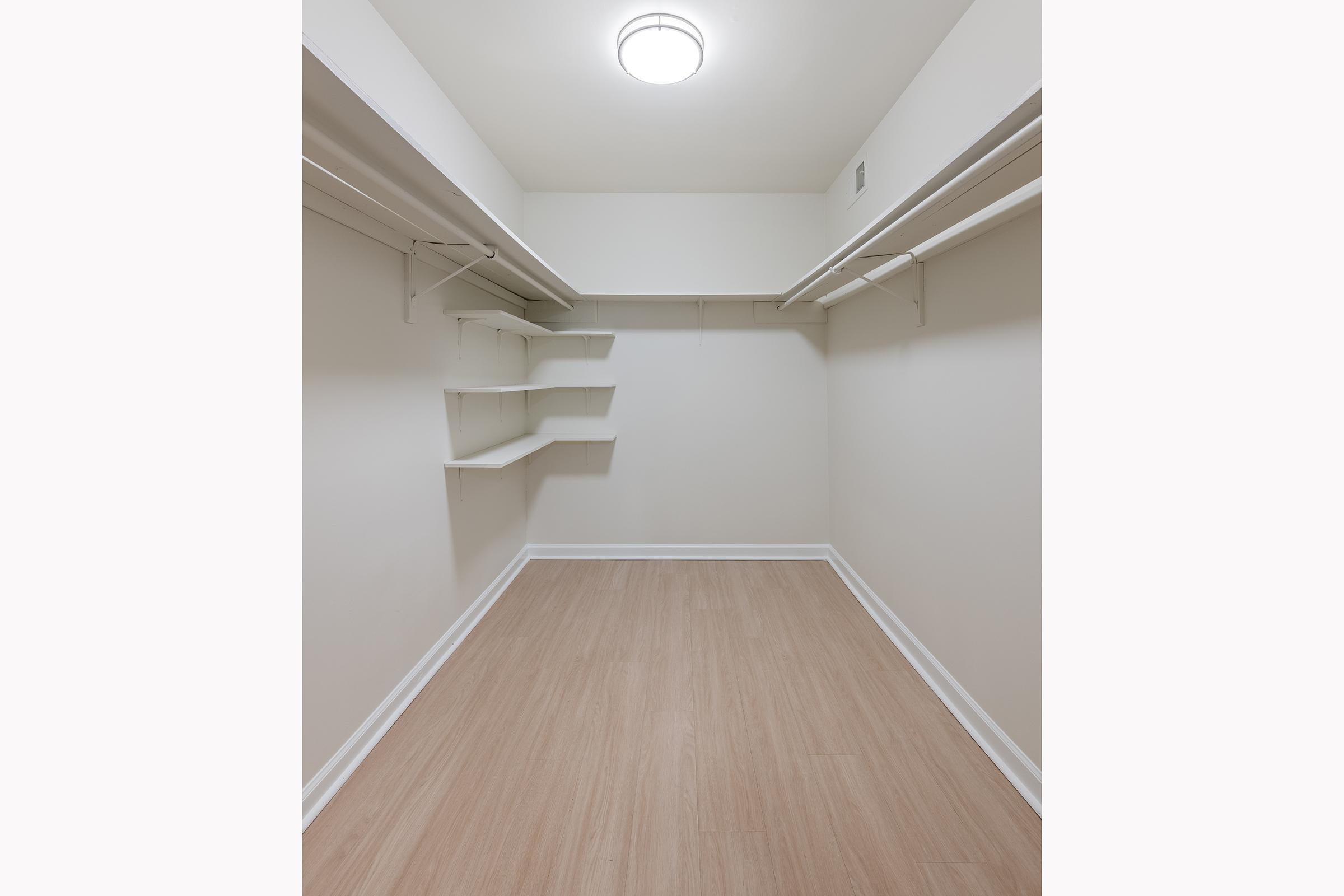 A spacious, empty closet with light-colored walls, wooden flooring, and a ceiling light fixture. The closet features several hanging shelves along the left side and a few wall-mounted shelves on the right, creating a neat and organized storage space.
