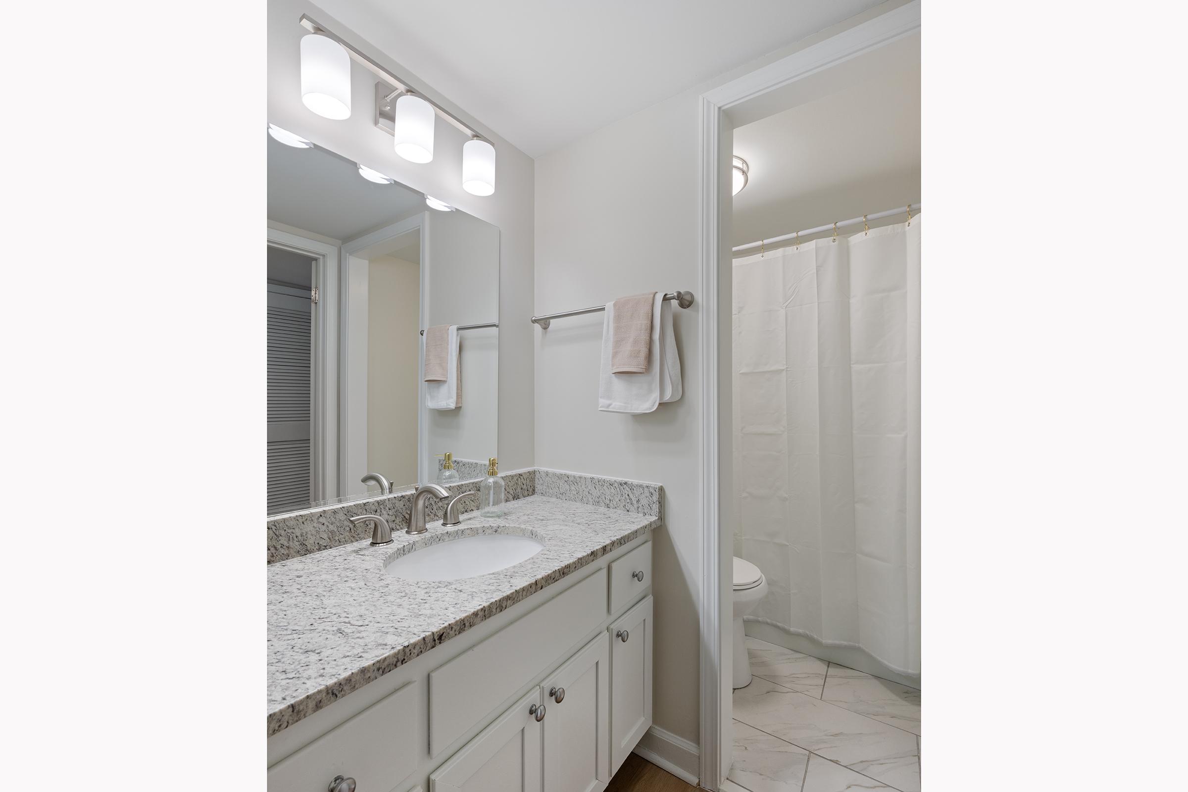 Modern bathroom with a light-colored granite countertop and double sink. Features a large mirror, stylish light fixtures, and white cabinetry. A white shower curtain partially obscures the shower area, which includes a toilet visible in the background. Soft beige towels hang on a towel rack.