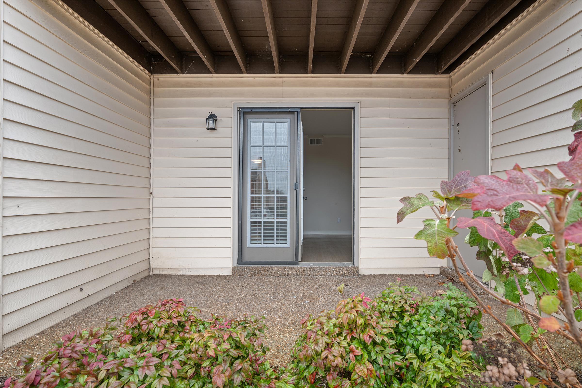 Front entrance of a home featuring a partially enclosed porch with a gray door and a window with vertical blinds. The walls are light-colored siding, and there are small decorative plants in the foreground. The area appears well-kept, inviting, and leads into an interior space visible through the door.