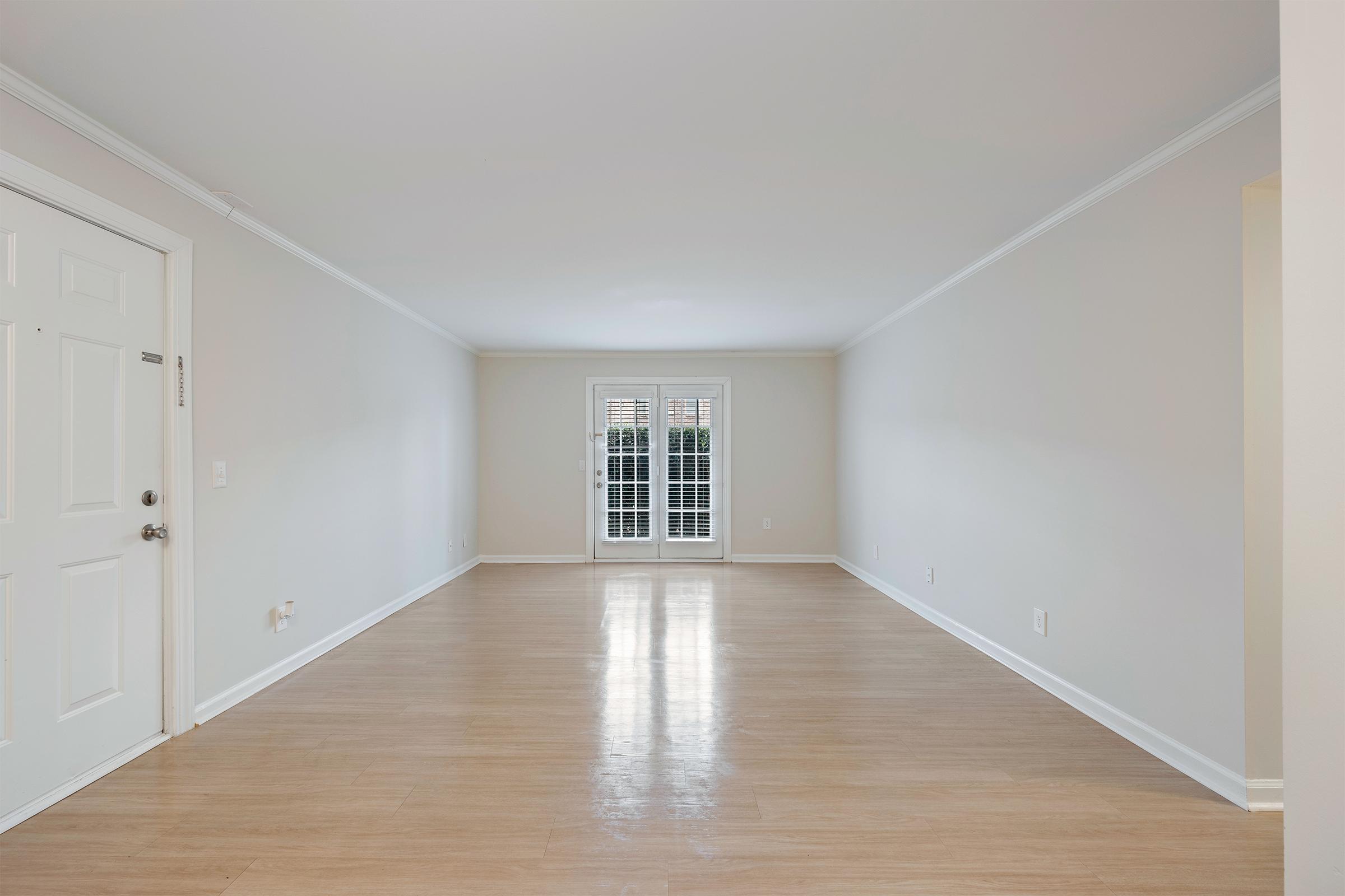 A spacious, empty living room featuring light-colored walls and a polished wooden floor. There are double doors leading to an outdoor area, framed by white plantation shutters. The overall ambiance is bright and inviting, with ample natural light illuminating the space.