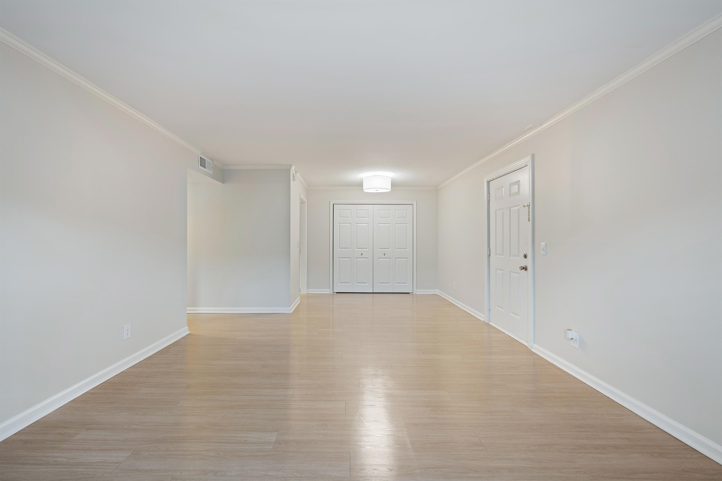 A spacious, empty room with light-colored walls and a smooth wooden floor. A ceiling light hangs in the center, illuminating the area. There are double doors with white panels on one side, and the overall layout is clean and minimalistic, creating a bright and airy atmosphere.