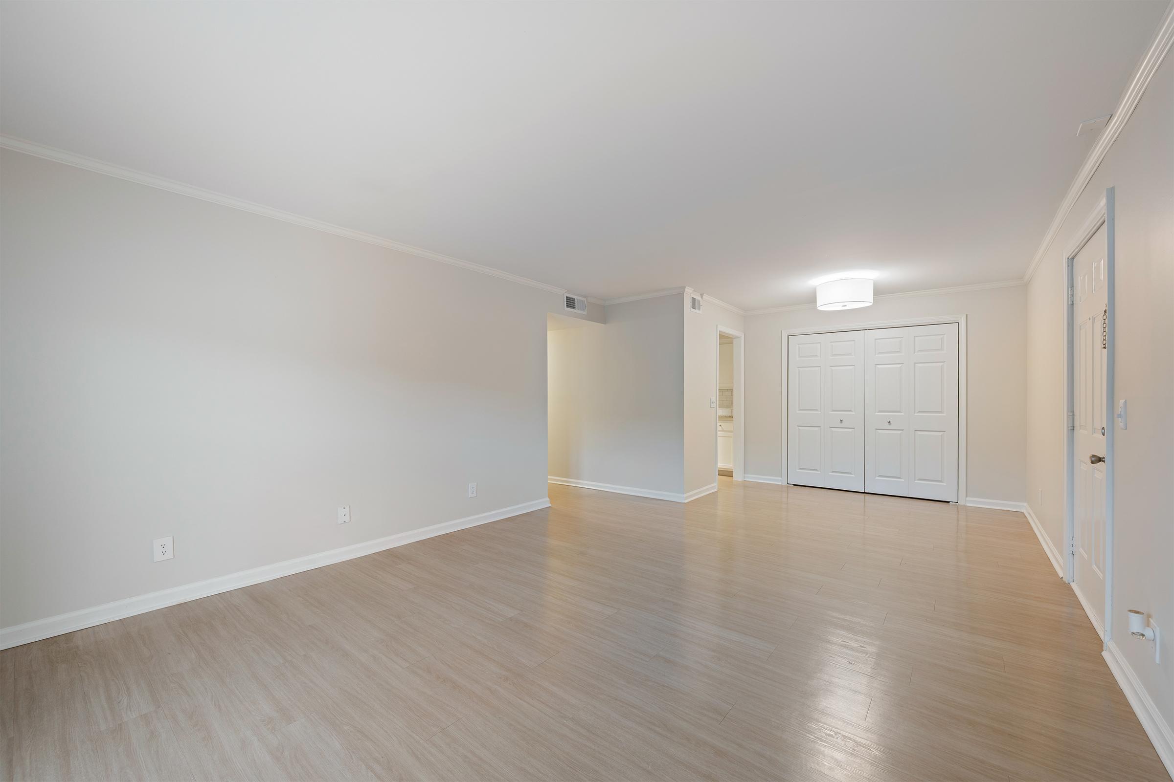 A spacious, empty living room with light-colored walls and flooring. A ceiling light fixture is visible, and the room features a doorway leading to another space. The closet doors are closed, and the overall atmosphere is bright and uncluttered, providing a clean and inviting environment.